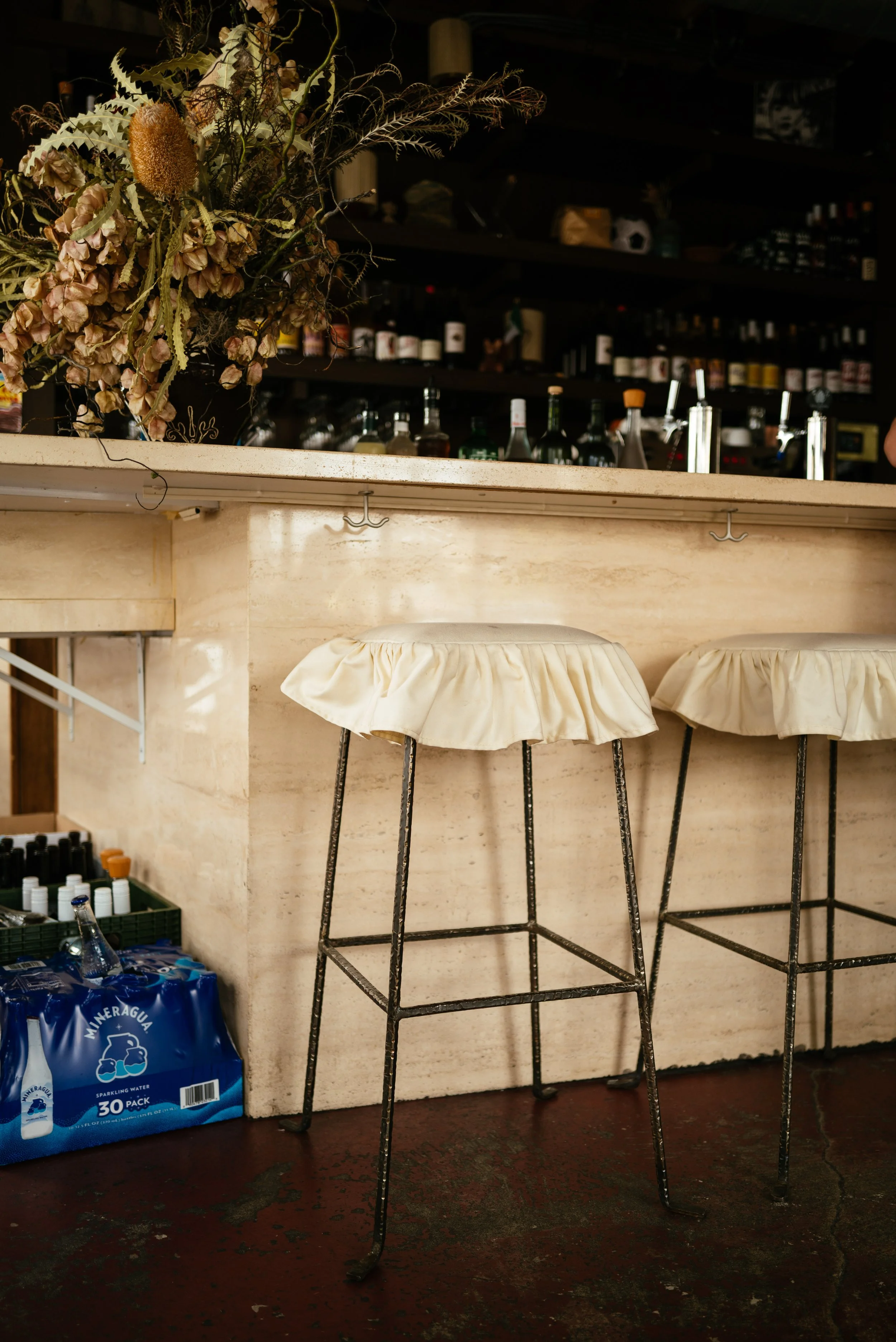 An empty bar counter with three tall bar stools with fabric seats, a large dried flower arrangement on the left, and shelves with bottles and supplies in the background.