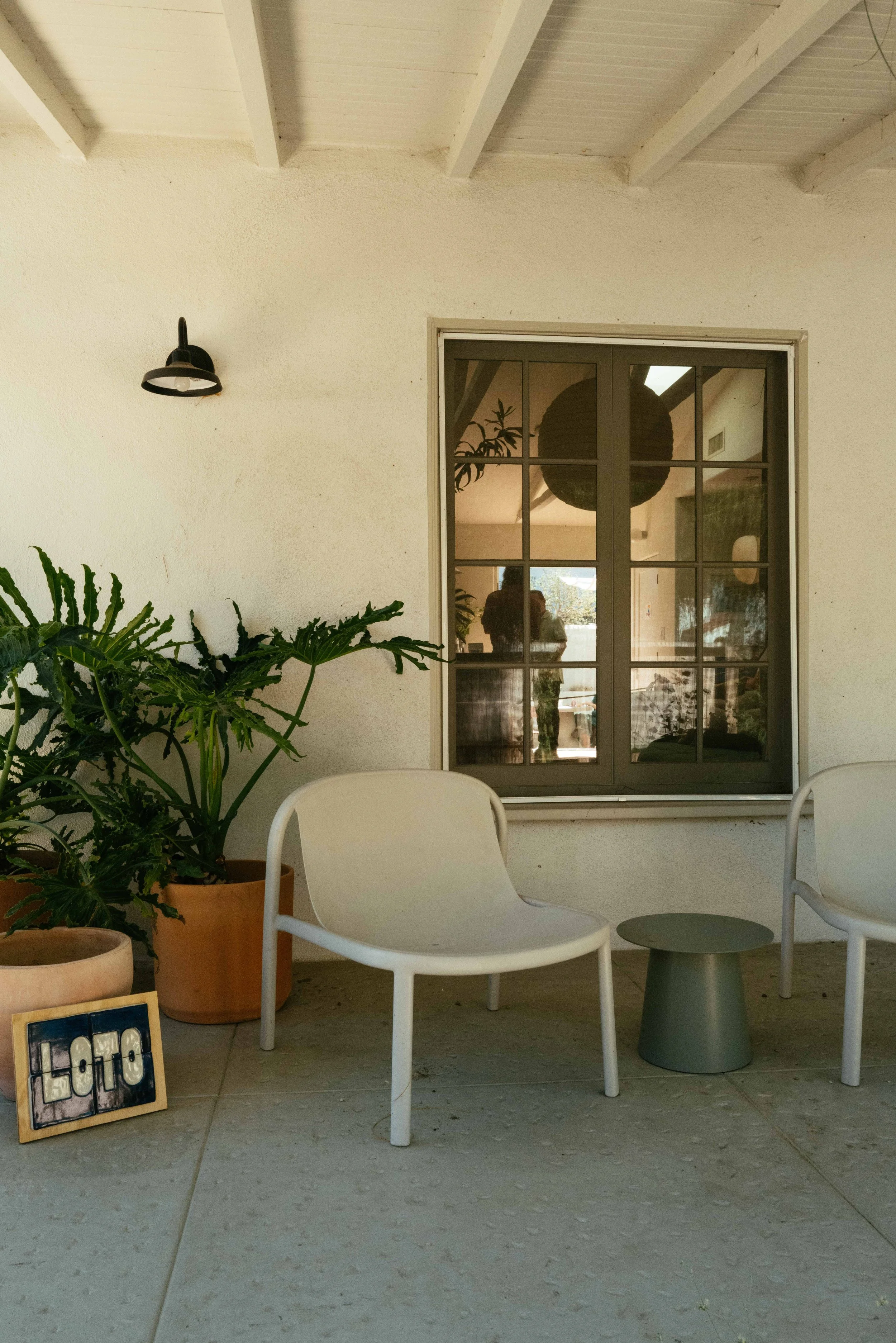 A patio scene with a white chair, a small green side table, and potted plants. There is a window with a reflection showing a person taking the photo. A sign near the plants reads 'LOFT4'.