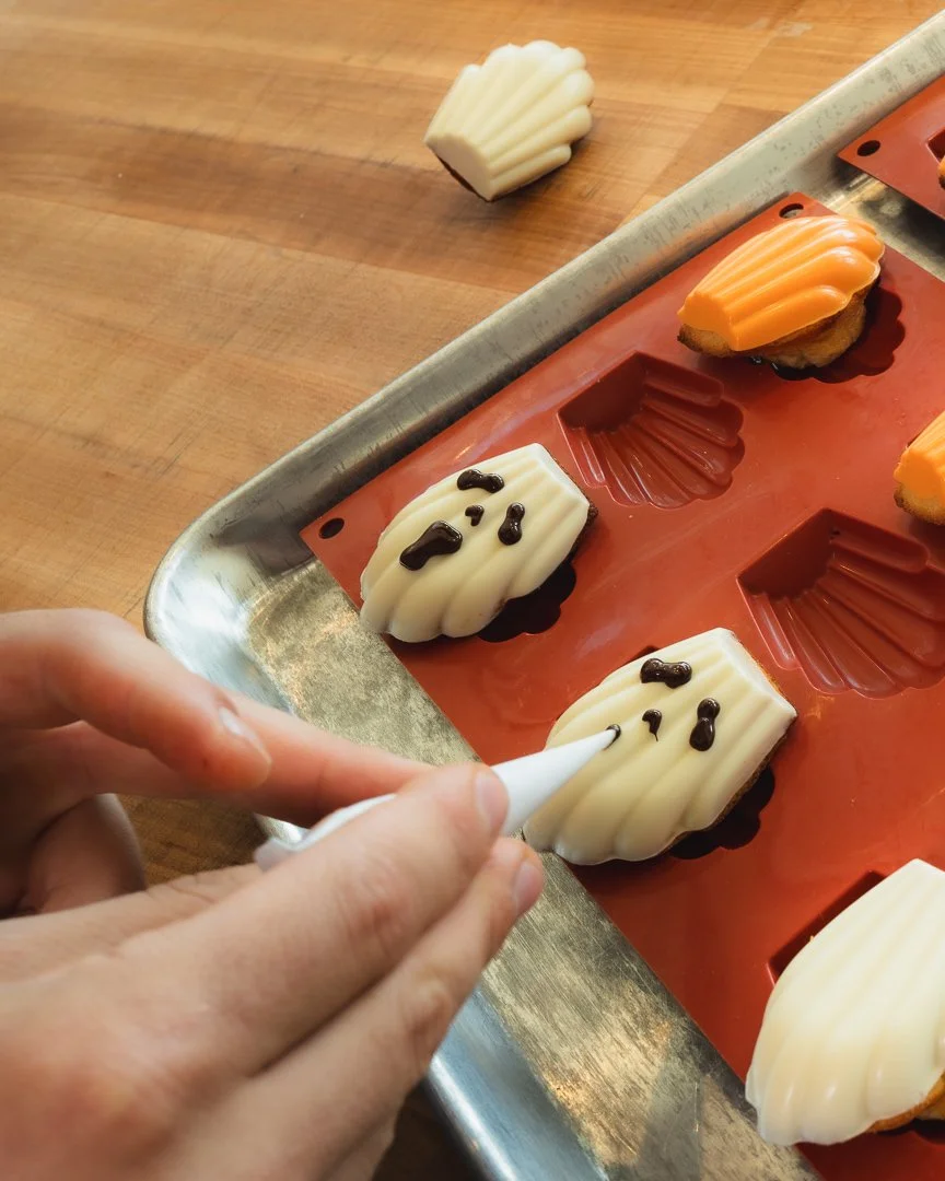 Baker piping chocolate faces onto Halloween madeleines, Louma Bakery