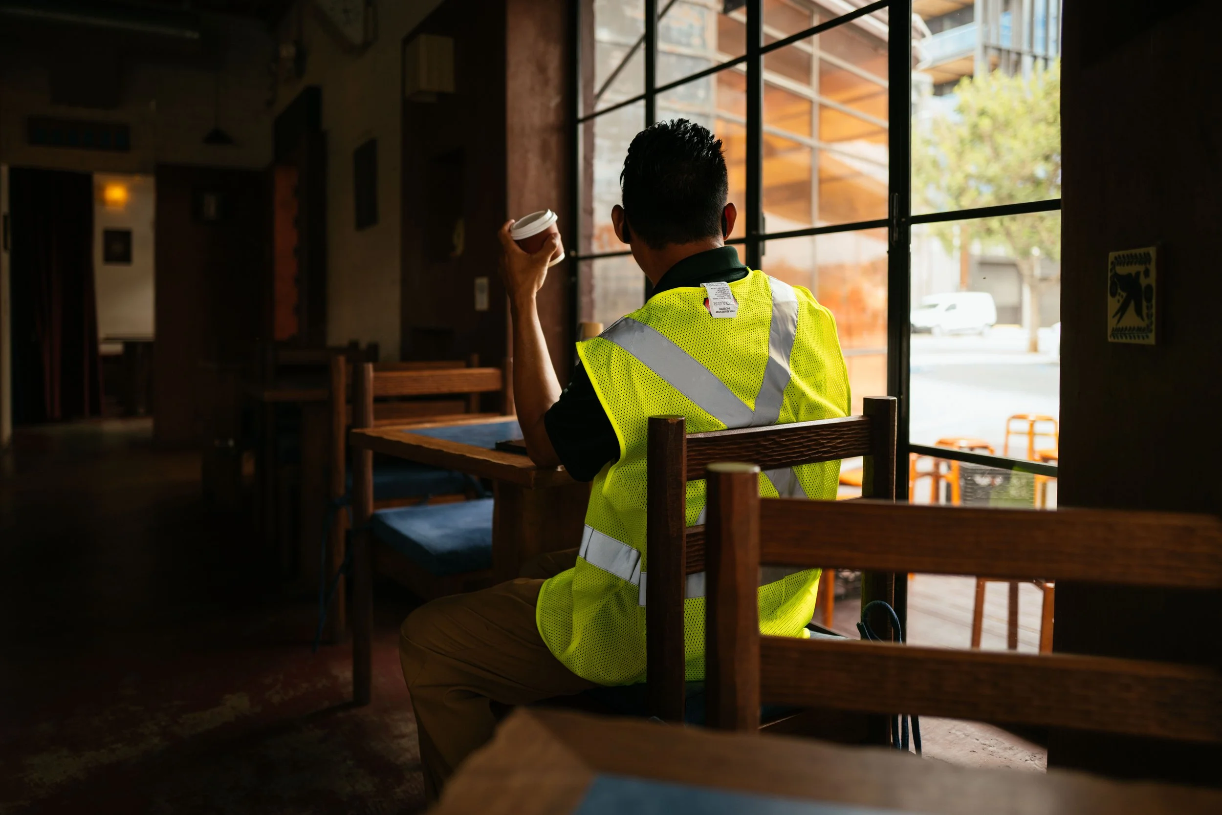 A man wearing a high-visibility yellow safety vest and khaki pants is sitting at a wooden table inside a cafe, holding a small disposable coffee cup. He is facing away from the camera, looking towards the large window that reveals a street scene outs