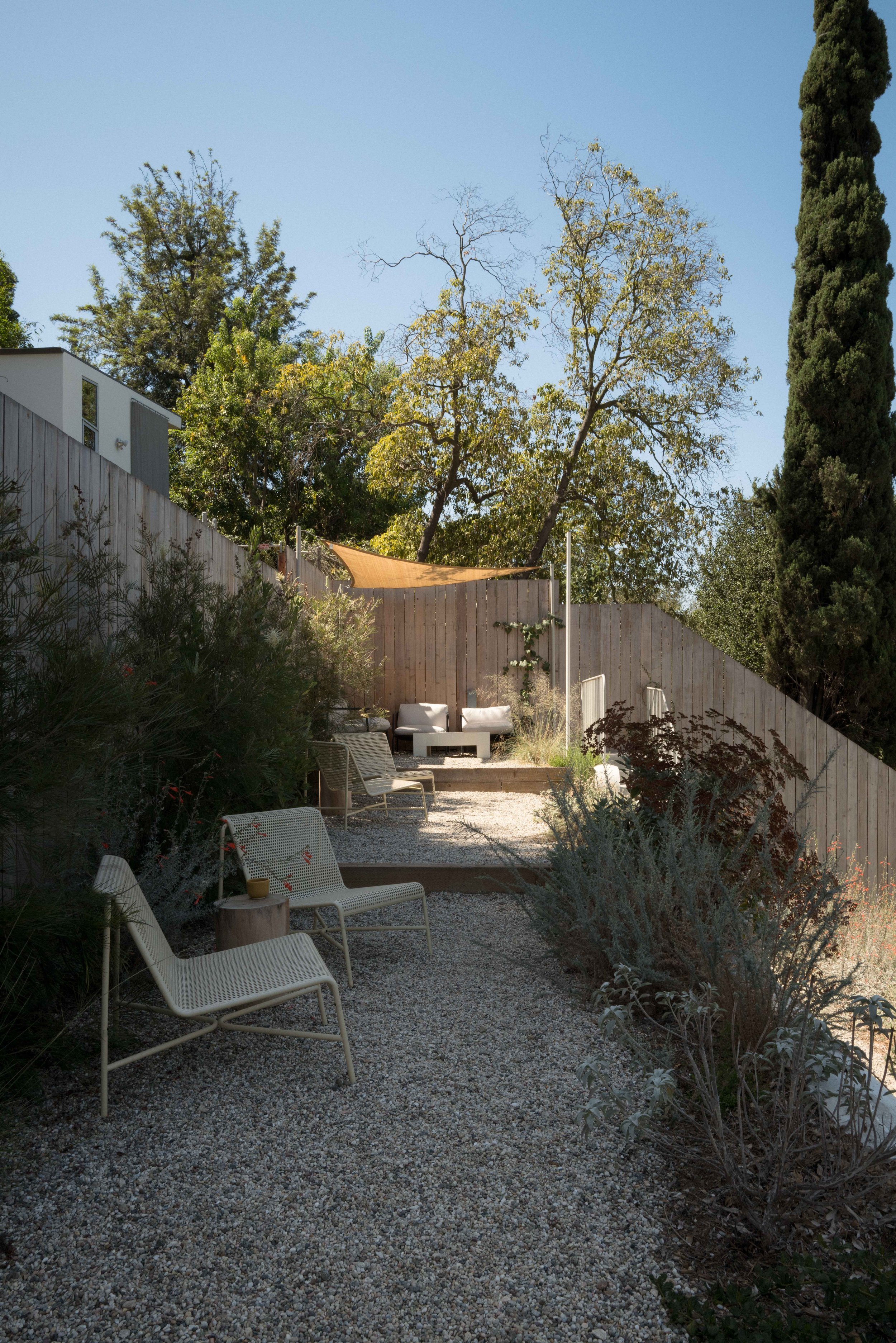 Outdoor backyard patio with white modern chairs, trees, and a wooden fence under a clear blue sky.