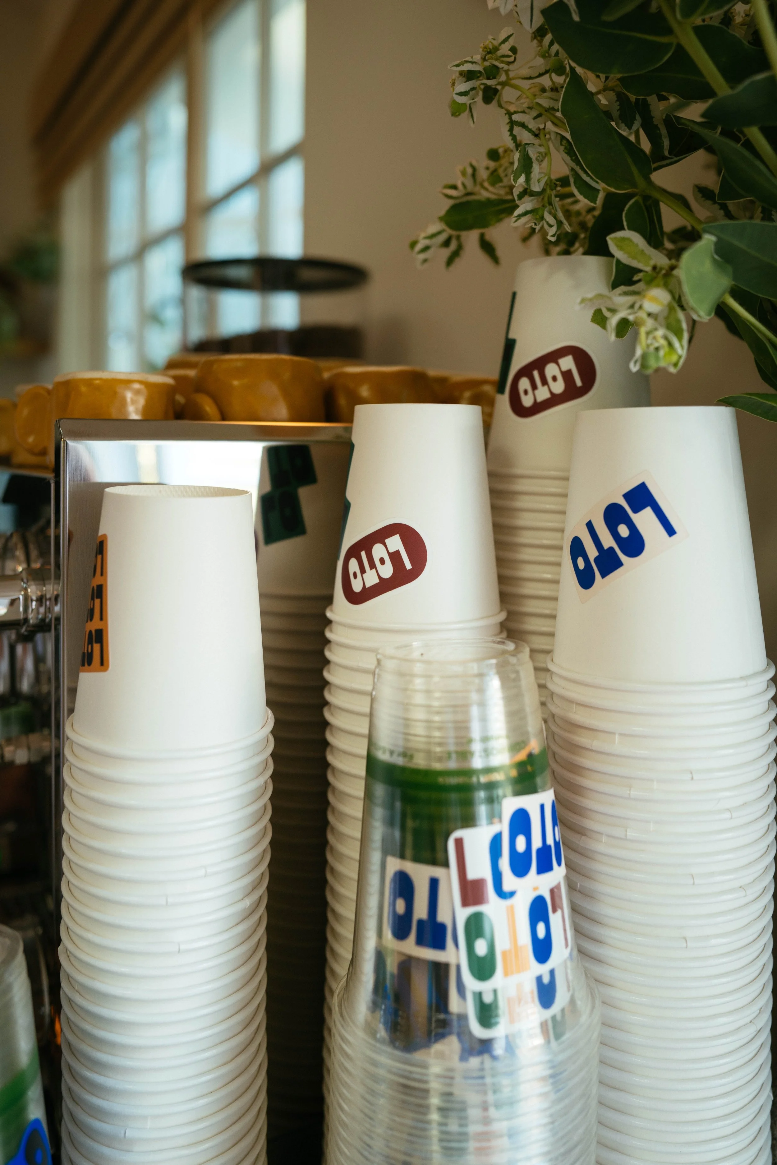 Stacked white disposable cups with colorful stickers and a glass water bottle in front, in a kitchen or café setting with green plants and sunlight streaming through window.