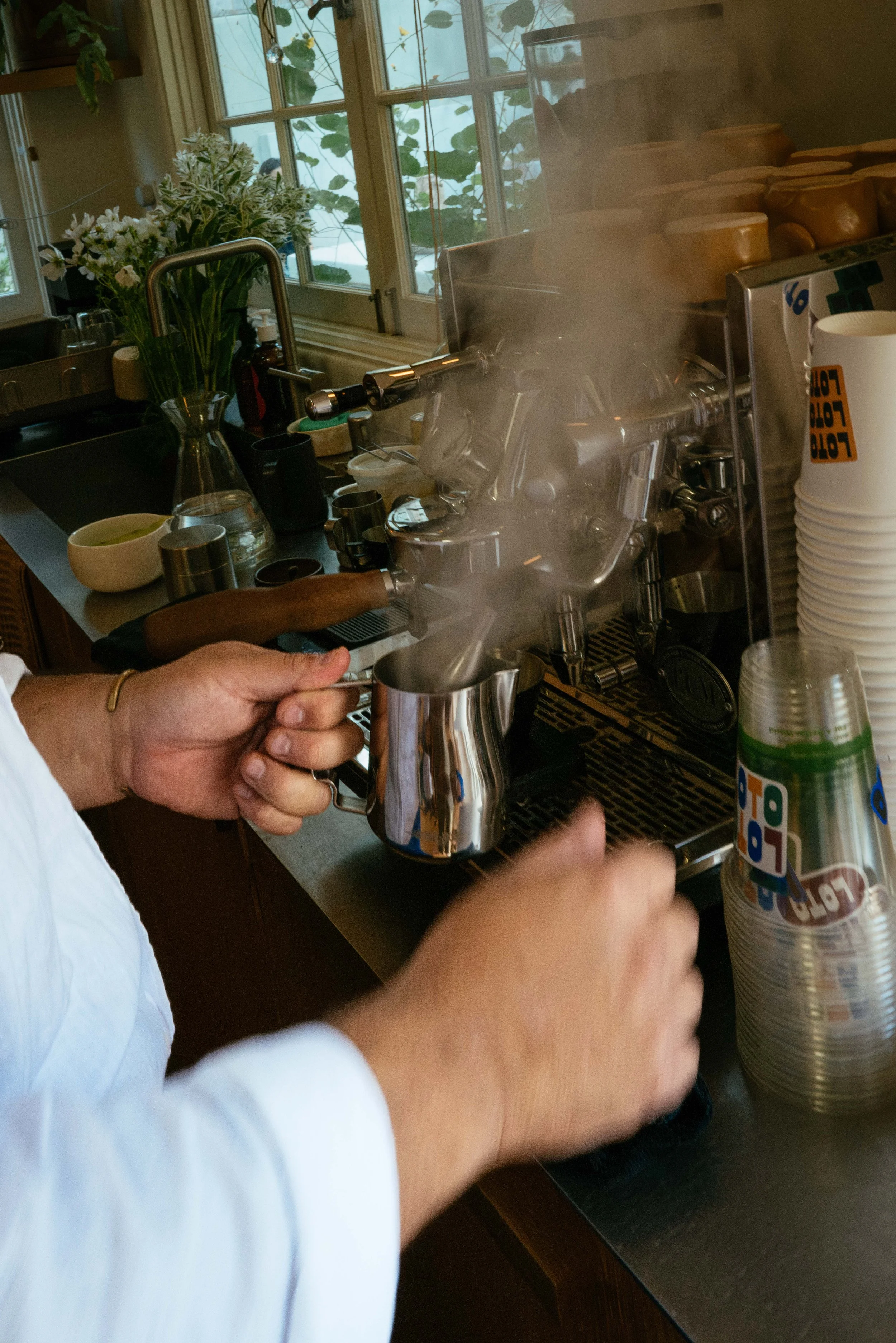 A person holding a steaming metal vessel under a coffee machine at a kitchen counter with a window and flower vase in the background.