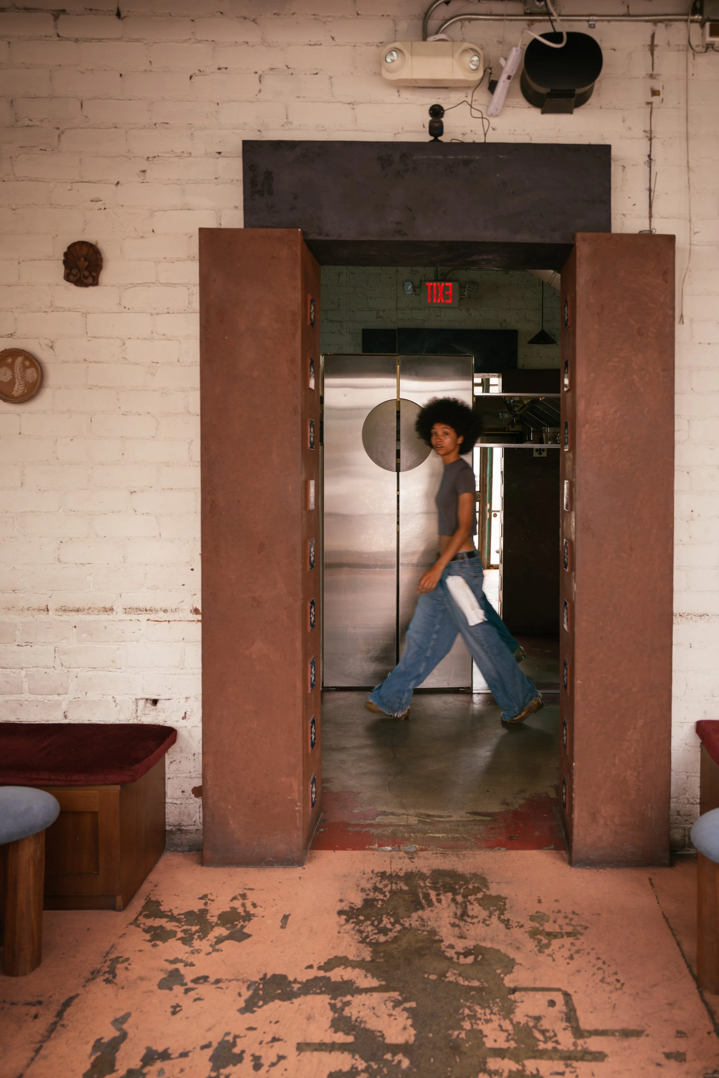 A woman with an afro hairstyle walking through an industrial-style space, passing through a brick archway with rust-colored walls, metal door, and vintage decor on the walls, with a distressed concrete floor.