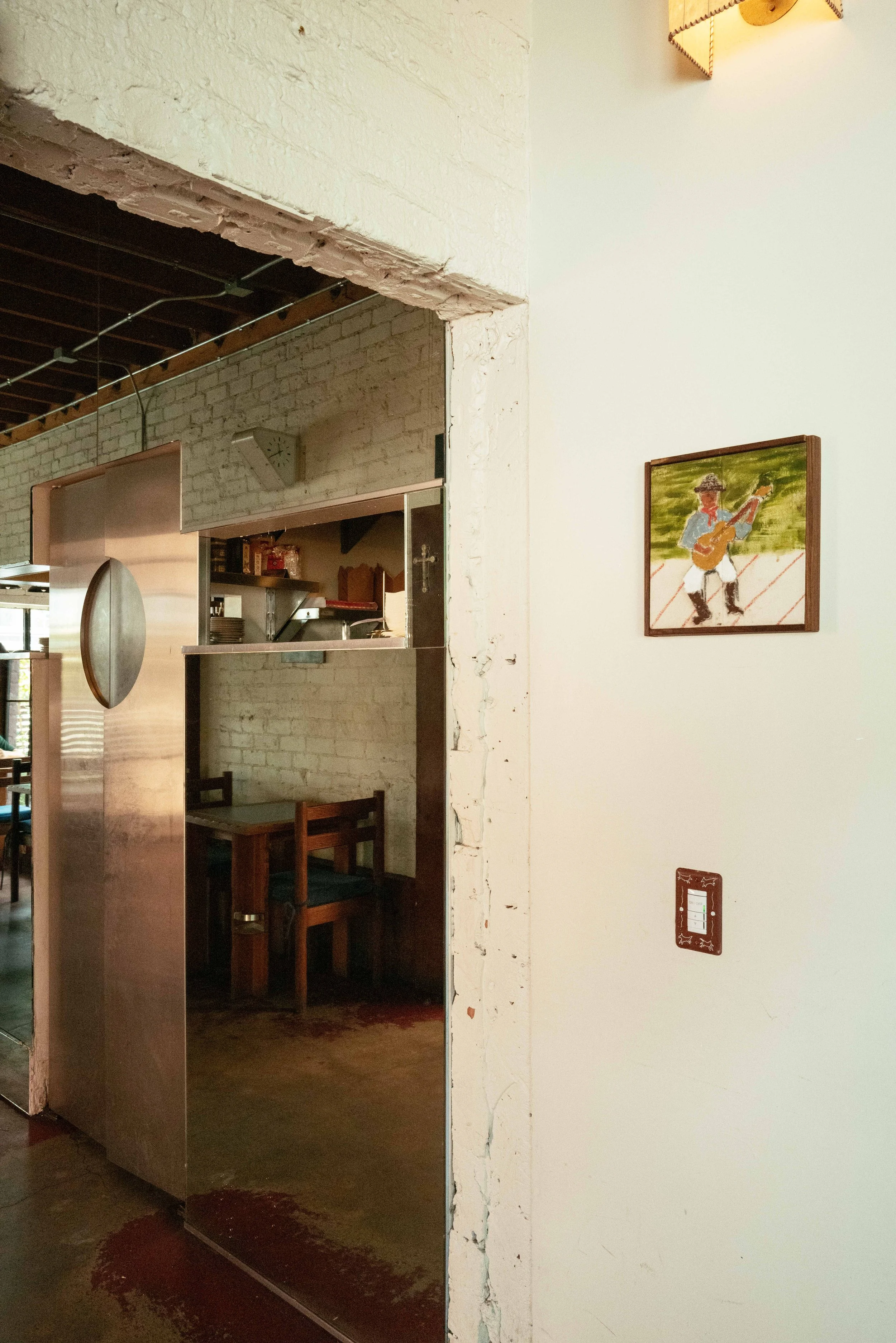 Interior of a restaurant or café with a brick wall, and a white painted wall with a painting of a boy playing guitar. There is a stainless steel door partially opened revealing tables and chairs inside. A small clock is on the brick wall, and a light
