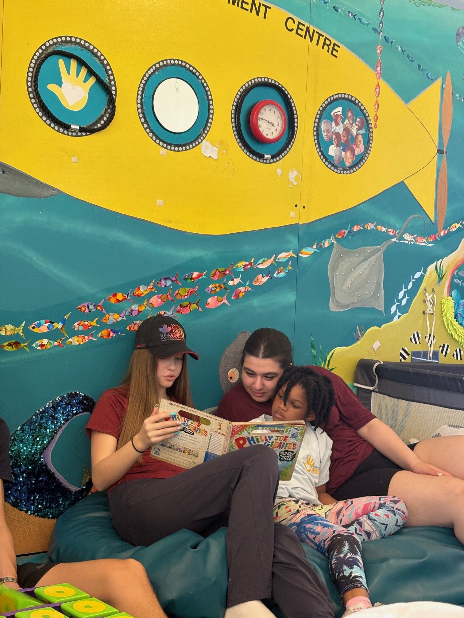 Group of young people sitting on a bean bag, one girl reading a colorful children's book, two girls leaning in and looking at the book, colorful wall with fish and ocean-themed decorations in the background.
