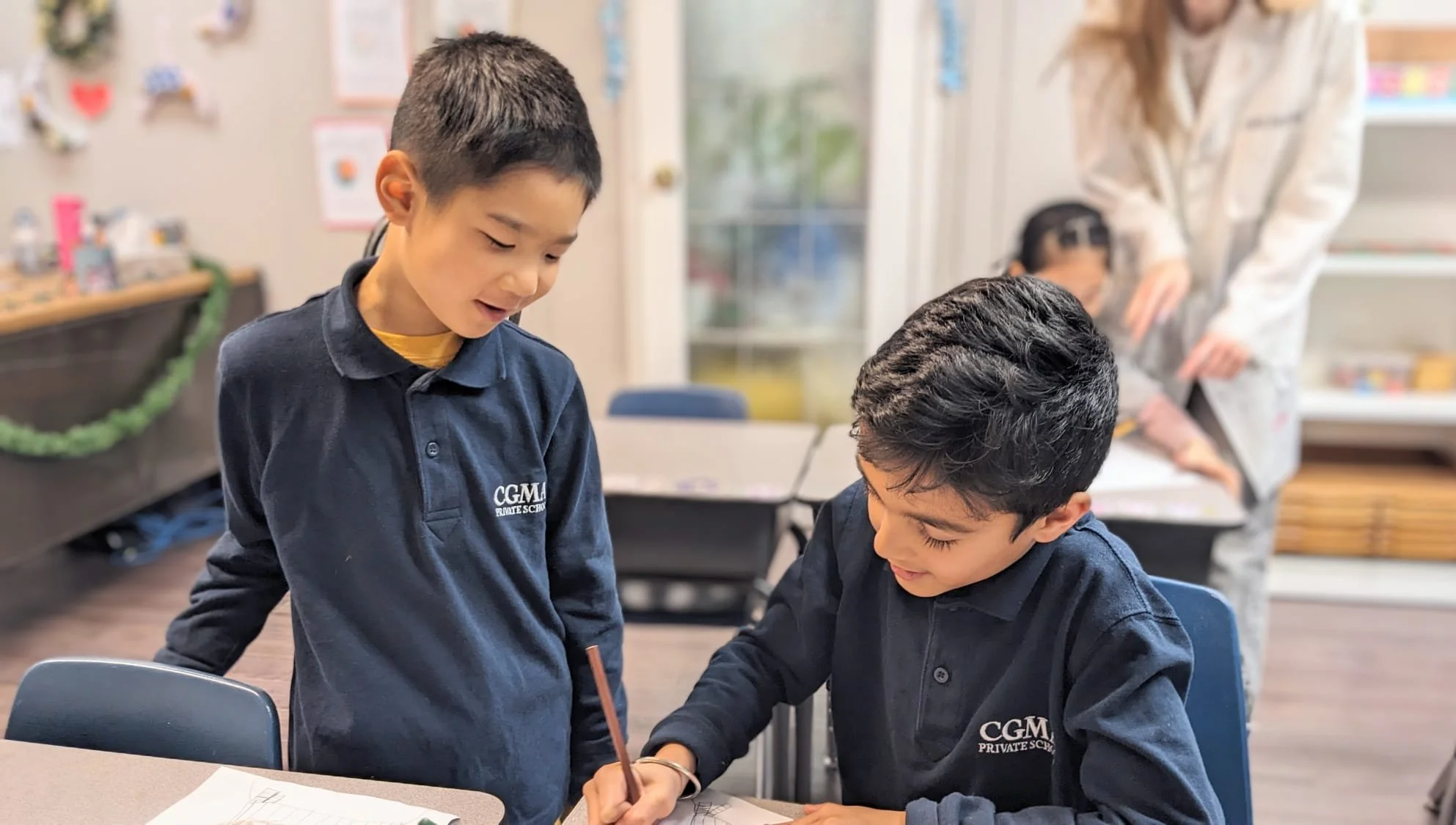 Two boys wearing navy blue school uniforms are working together at a table, with one standing and the other seated. They appear to be collaborating on a school assignment or project.