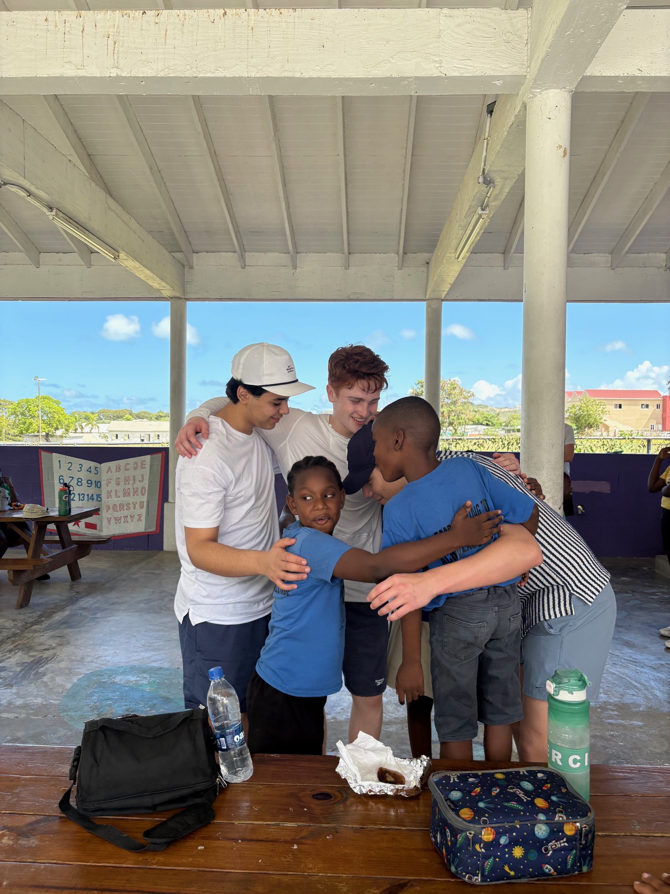 A group of children and teenagers hugging and smiling under a pavilion with a blue sky and green trees in the background. There is a table in front with a backpack, water bottle, and lunch items.