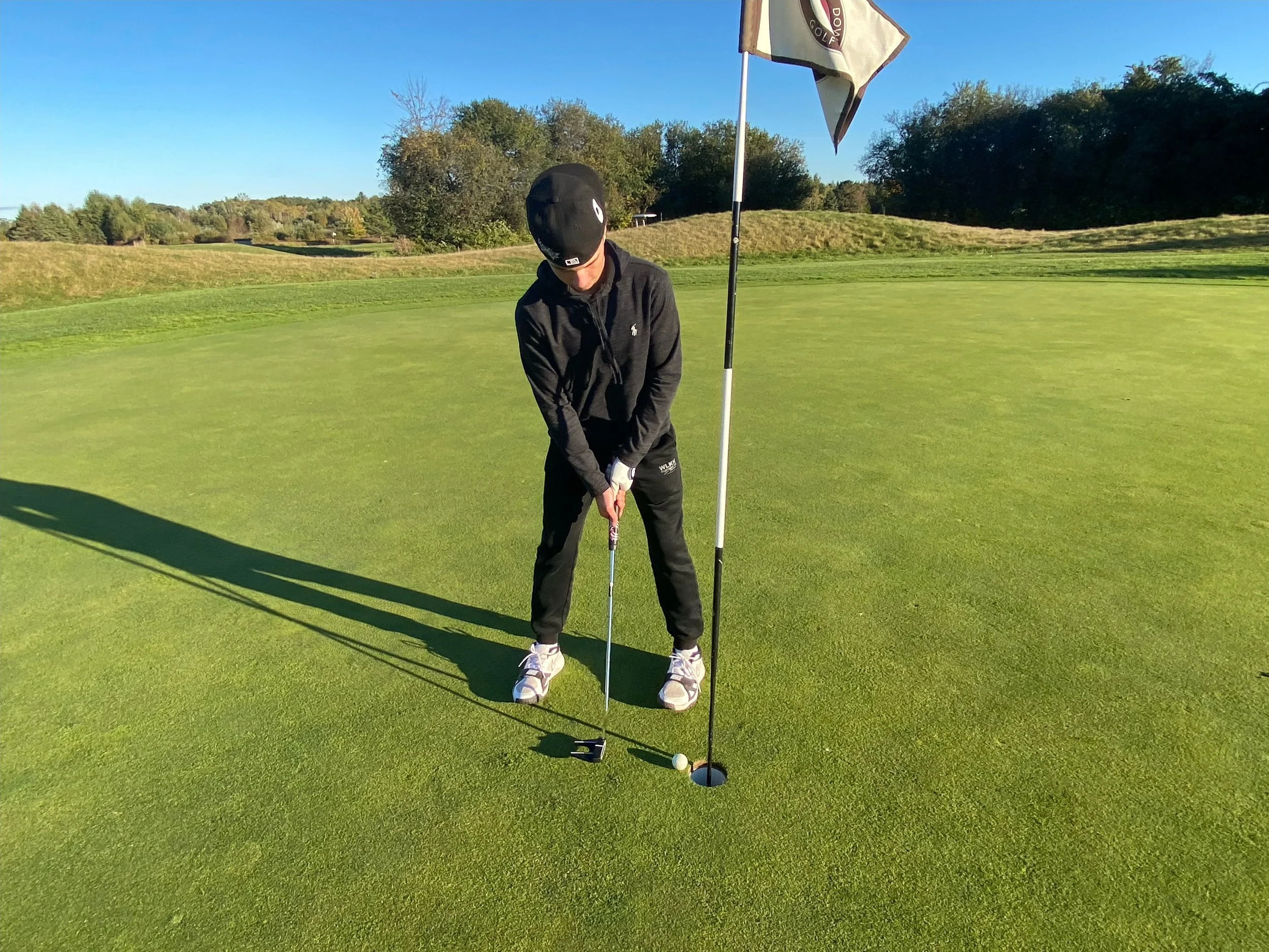 A man playing golf on a sunny day, standing near the hole on the green, holding a putter, with a golf flag behind him and trees in the background.