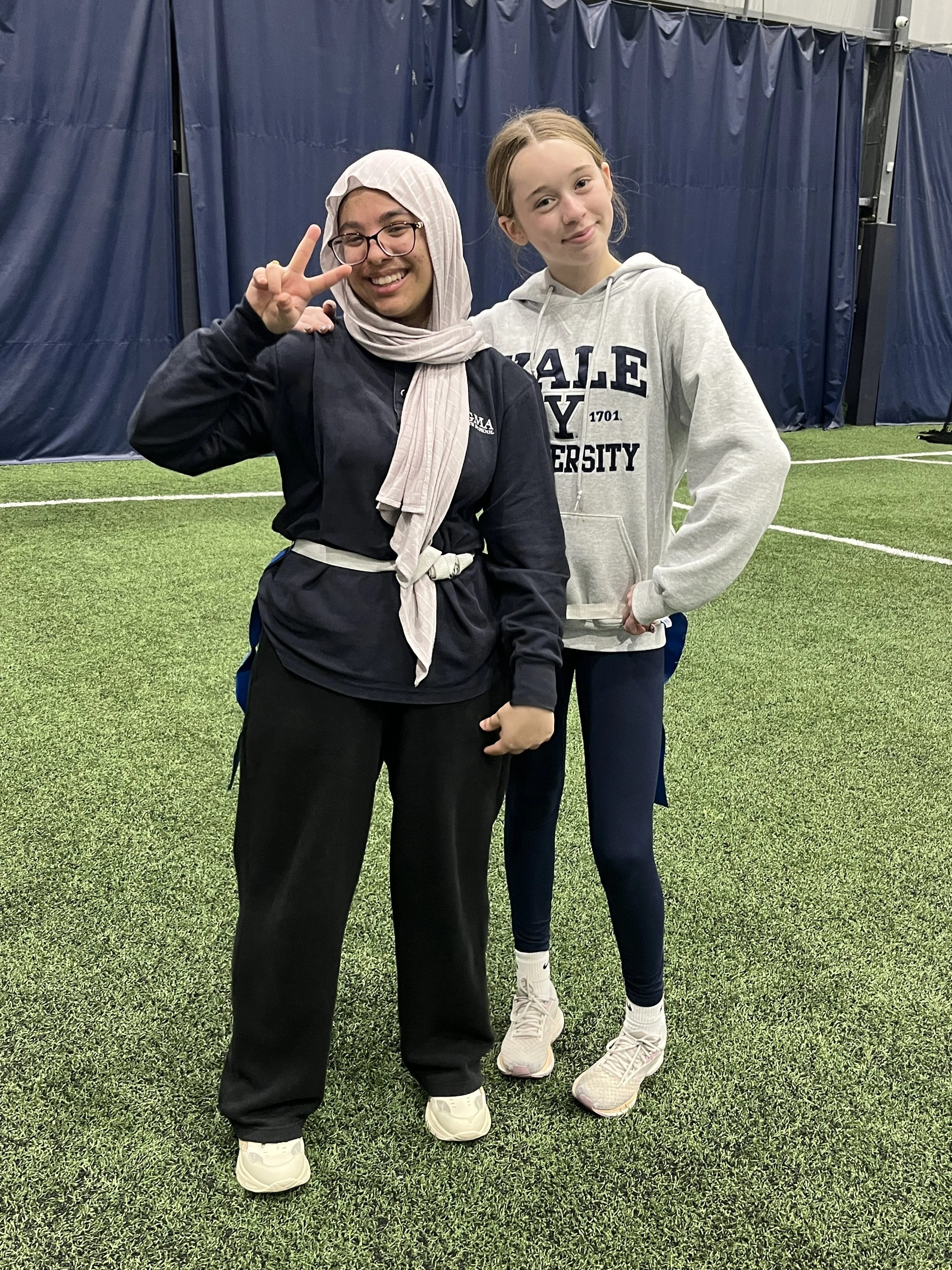 Two young women standing on indoor turf. One is wearing glasses, a hijab, a black jacket, and black pants, making a peace sign. The other is in a gray hoodie with Yale University written on it, navy blue leggings, and white sneakers, with her arm around the first woman.
