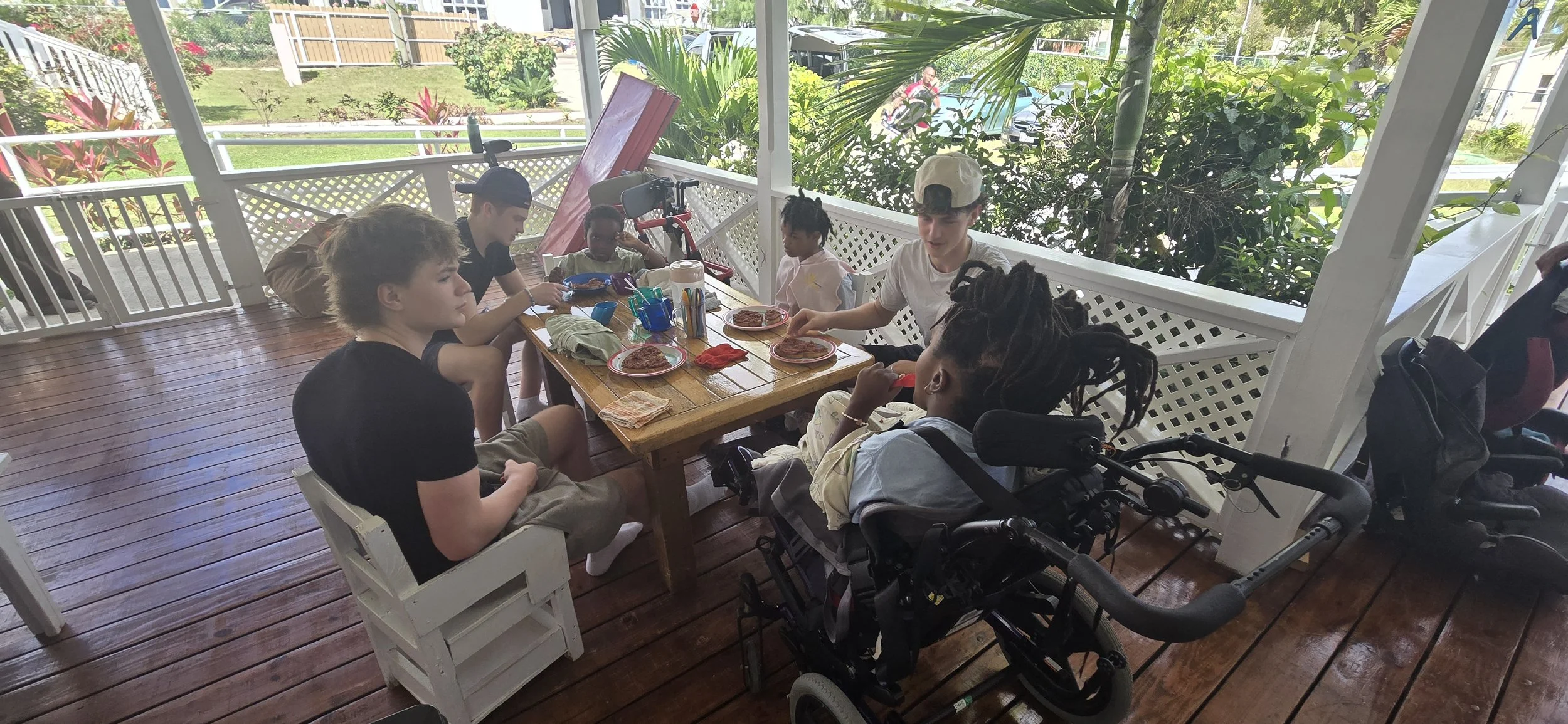 A diverse group of children and teenagers are sitting around a wooden table on a porch, enjoying a meal with plates of pizza. Some are engaged with their food, while others are talking or looking around. The porch has a decorative white railing and i
