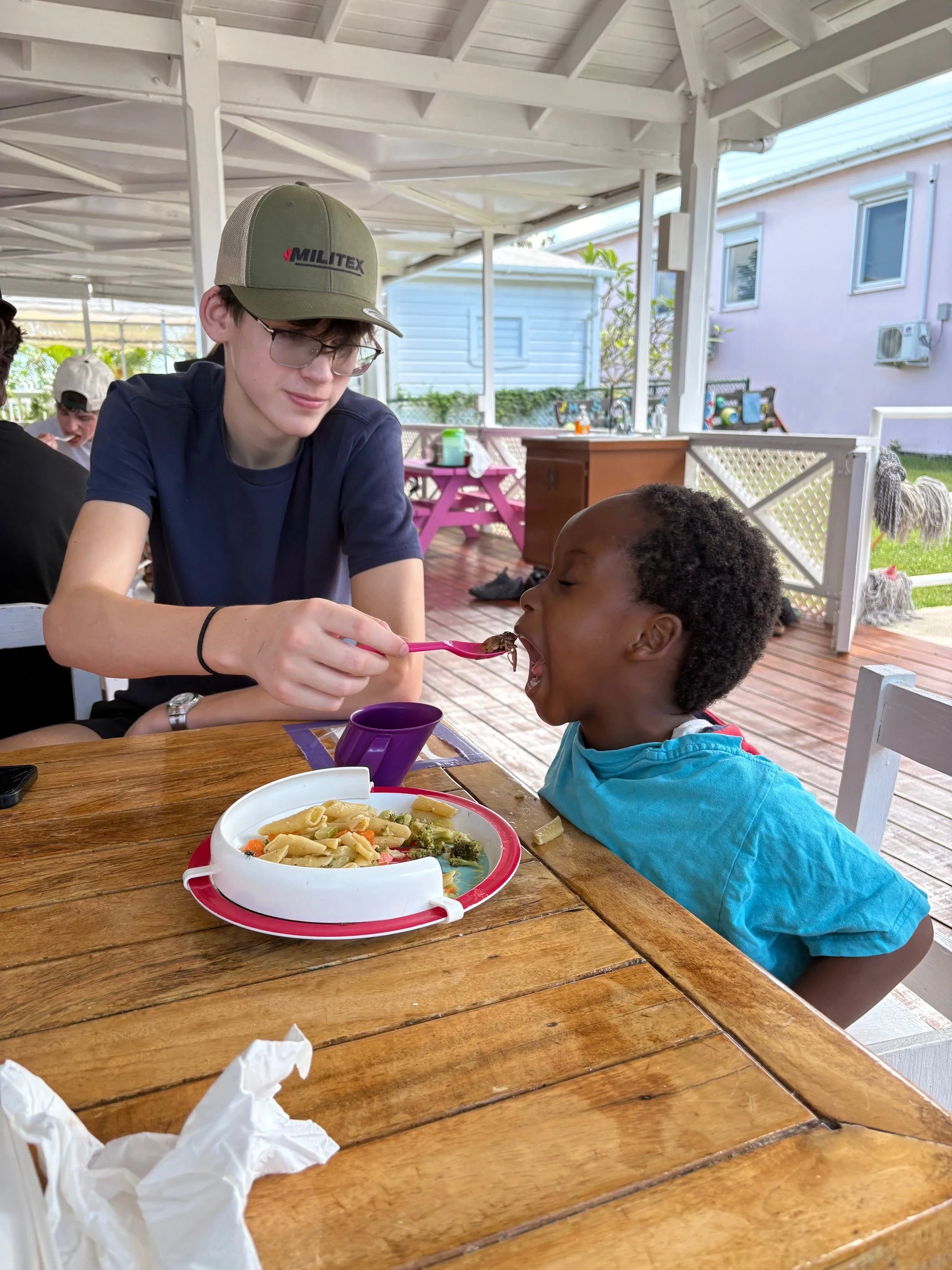 A young man with glasses wearing a green cap and a young boy in a blue shirt are sitting at a wooden table on a porch. The young man is feeding the boy with a fork. There is a plate of pasta and broccoli in front of them. The porch has a white roof a
