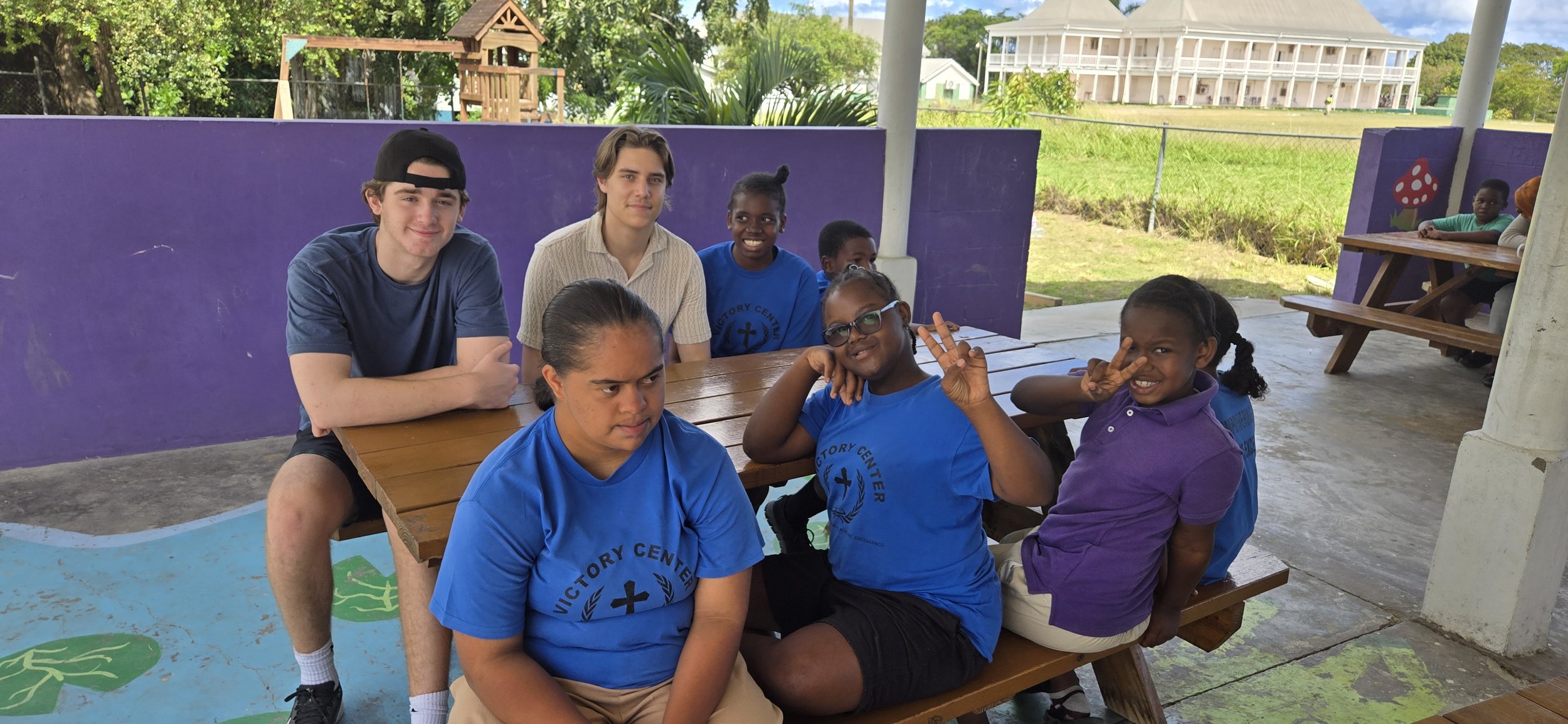 Group of children and teenagers sitting at a wooden picnic table, smiling and making peace signs, on an outdoor covered patio with purple walls, surrounded by green trees and a grassy field.