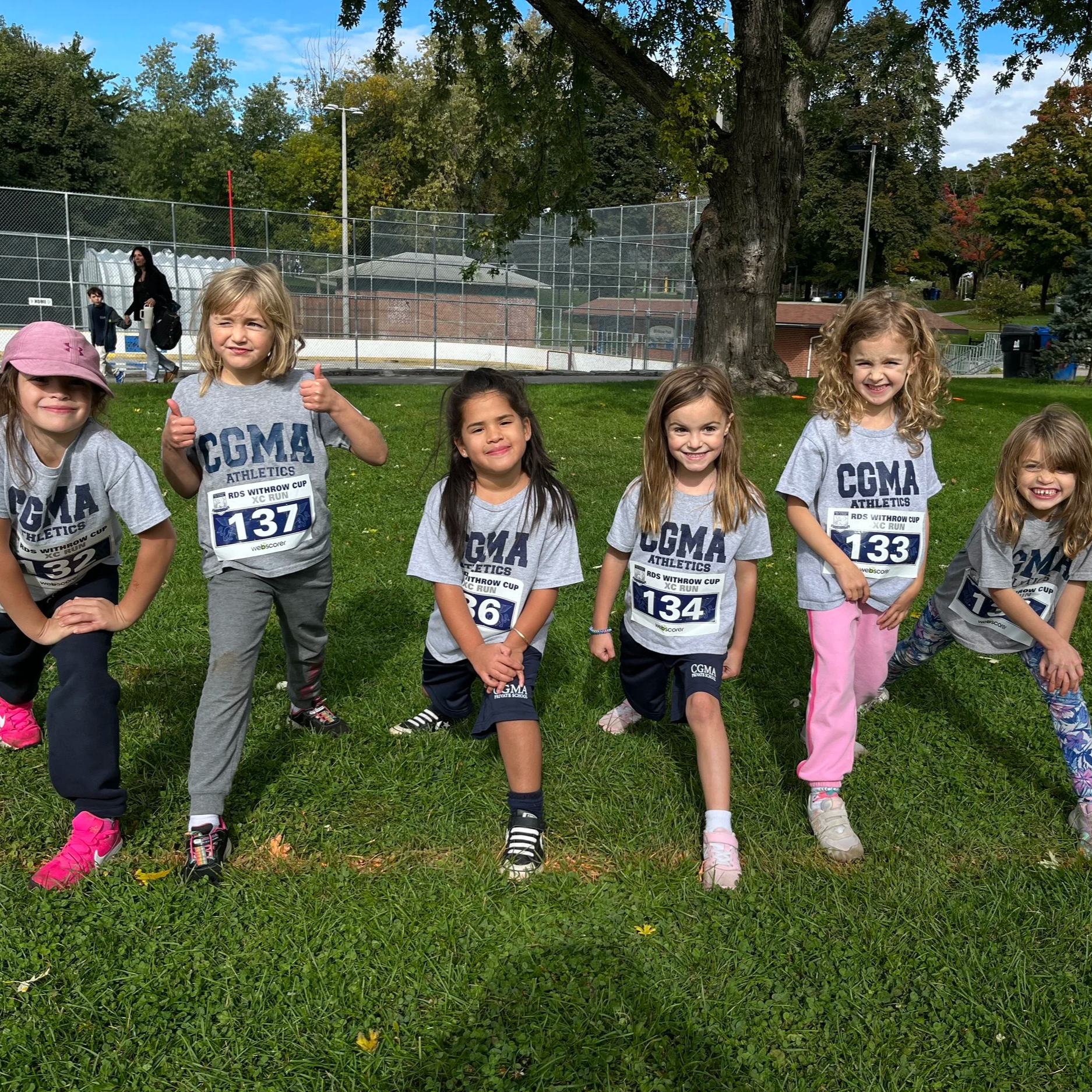 A group of young girls standing outdoors on grass, some giving thumbs up, wearing matching gray T-shirts with 'CGMA ATHLETICS' printed on them, and race bibs, with a fence and trees in the background.
