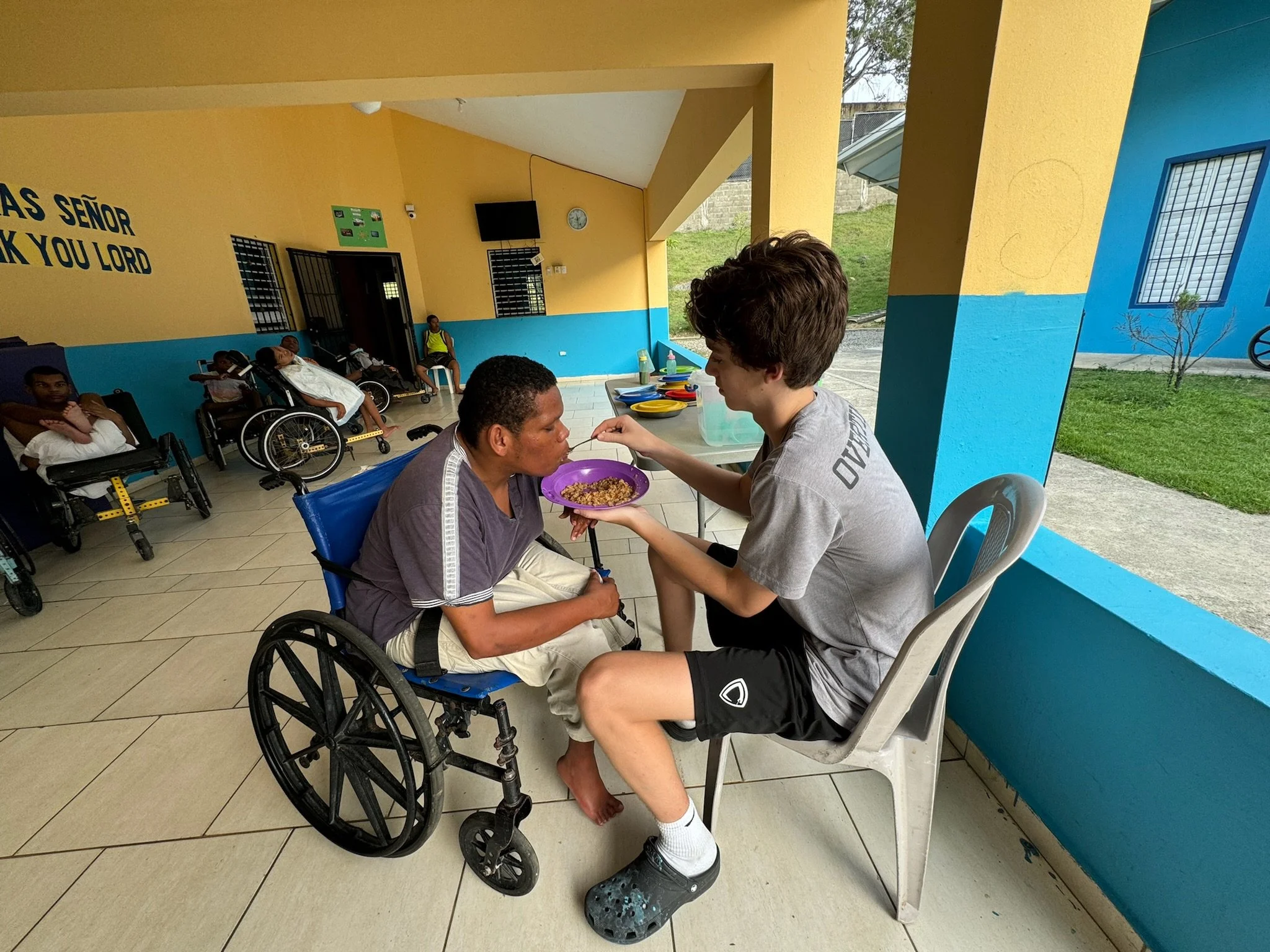 A young man feeding an elderly woman in a wheelchair at a community center or outdoor shelter, with other people in wheelchairs in the background. The area has yellow and blue walls, and outdoor scenery visible through an opening.