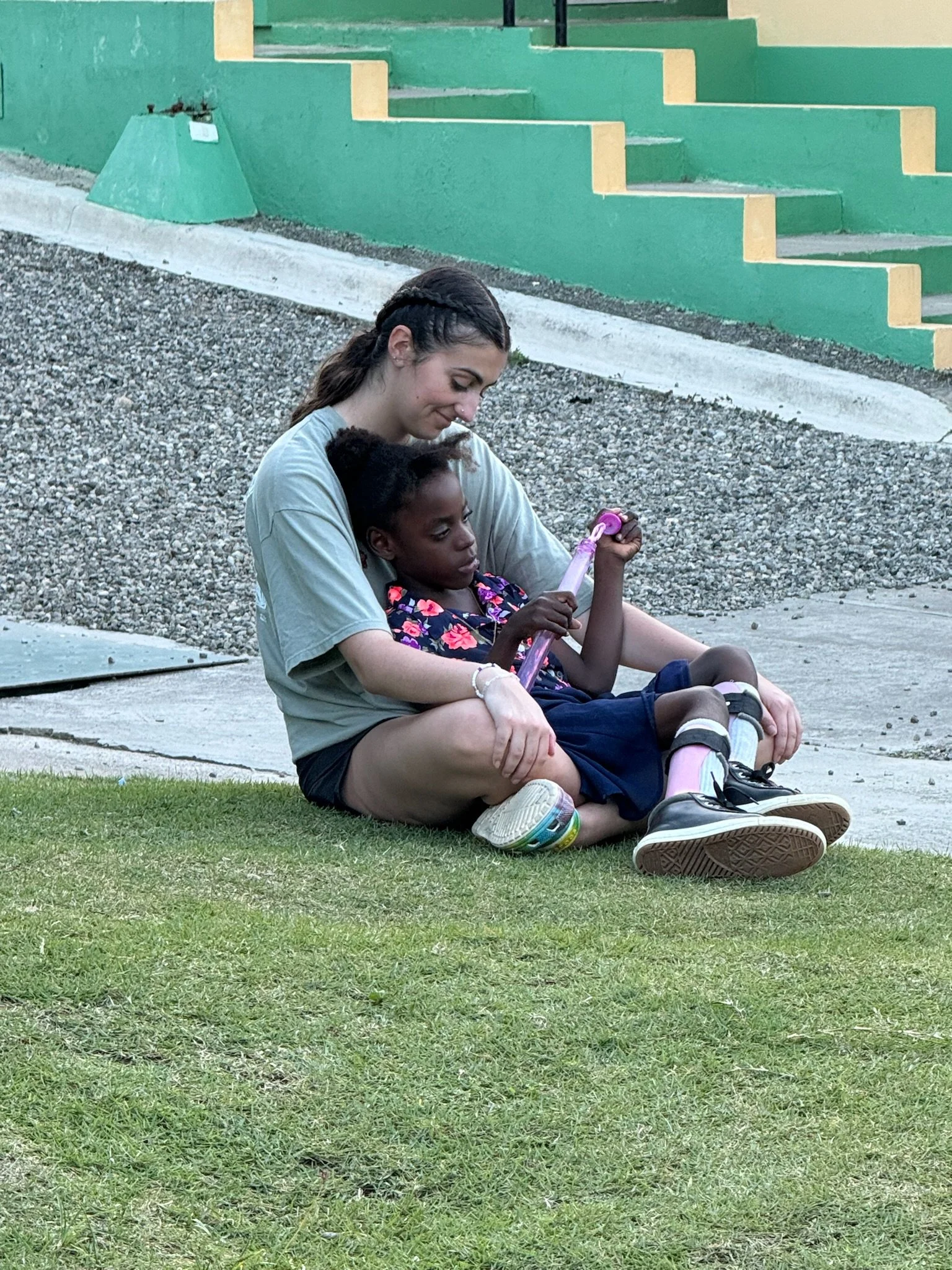 A woman sitting on the grass with a young girl, both smiling and looking at a pink toy the girl is holding. They are outdoors near a building with green and yellow steps.