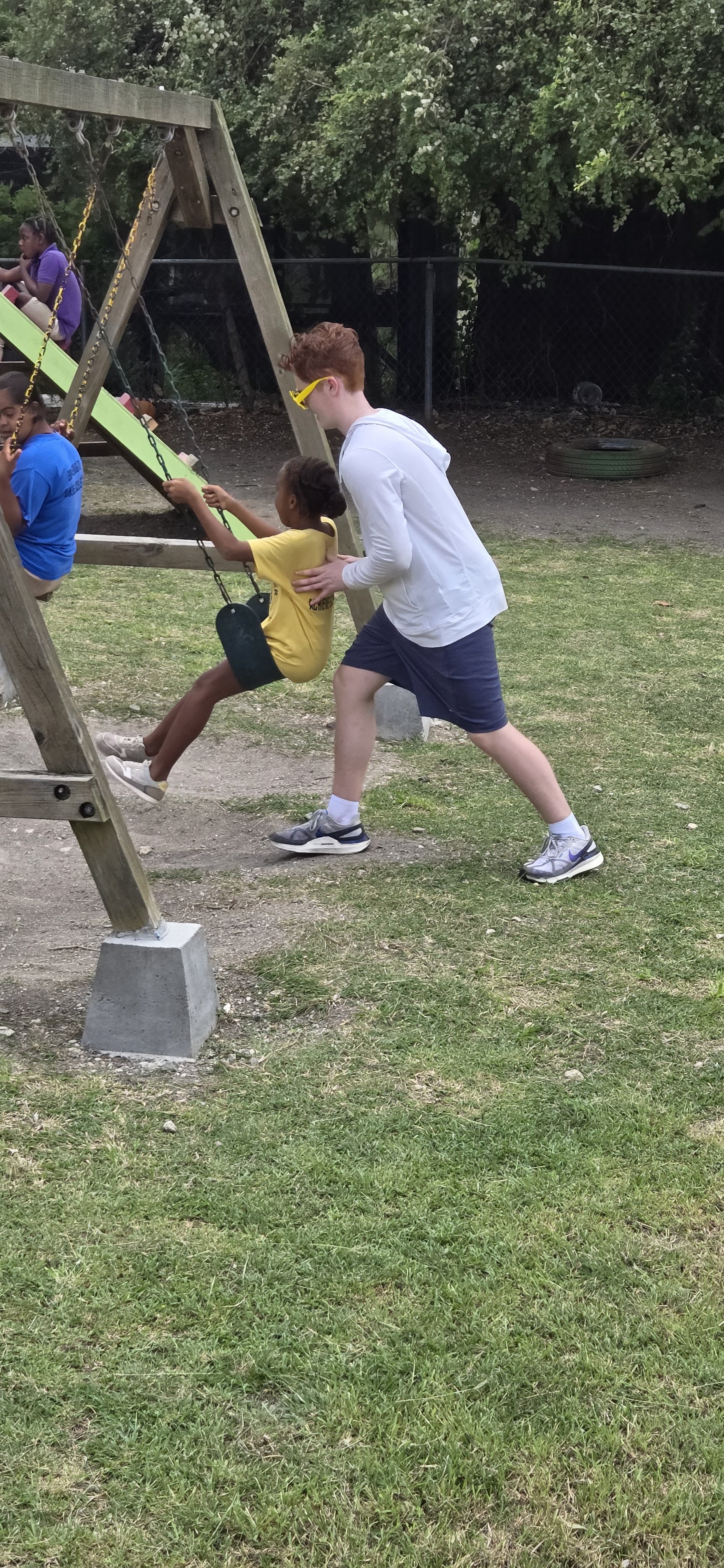 A young boy in a white hoodie and shorts pushing a girl on a swing at a playground. The girl is wearing a yellow shirt and black shorts. Other children are visible on the swings in the background.