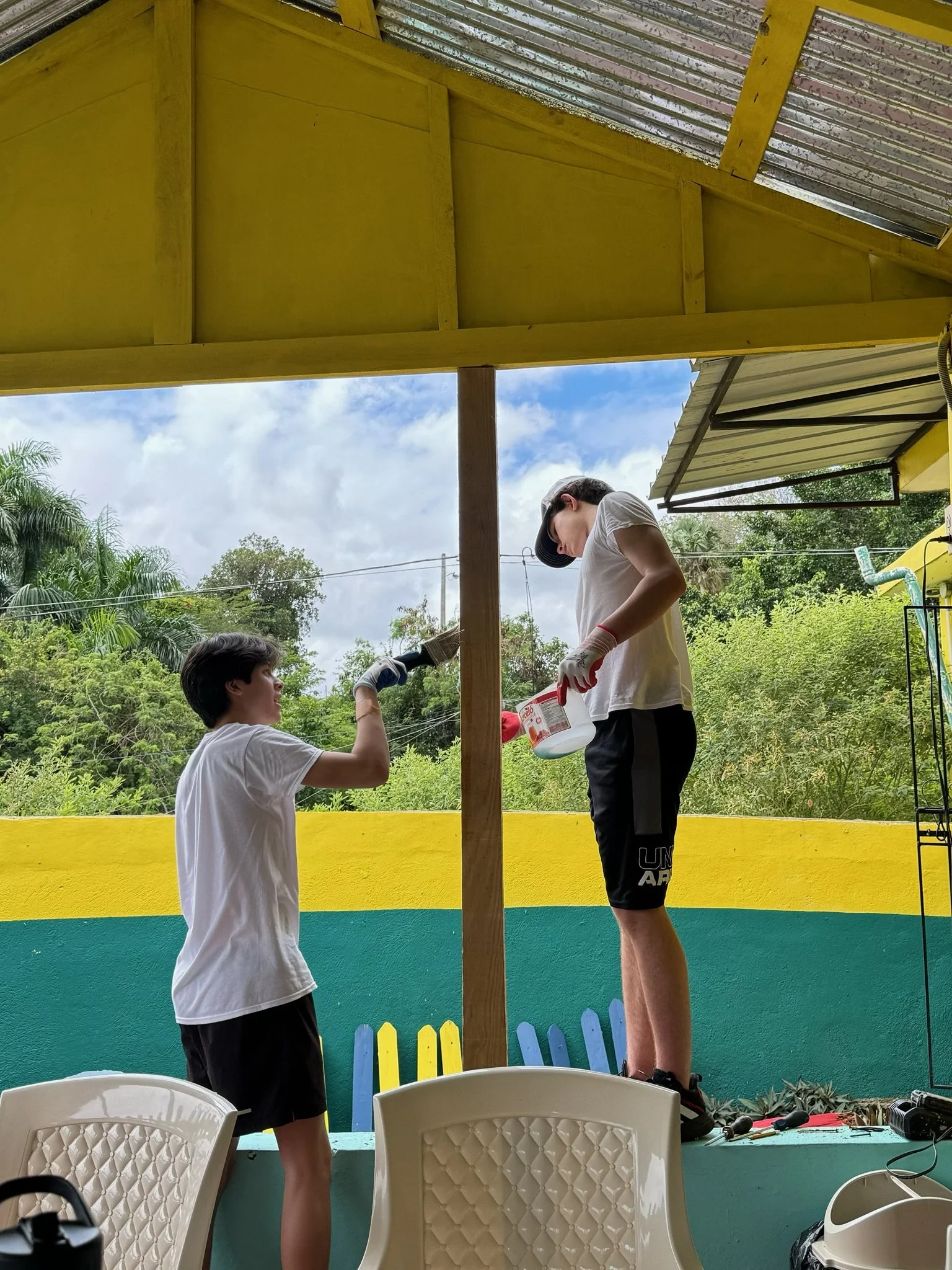 Two boys, one standing on a platform and the other below, working on a wooden structure outdoors surrounded by trees, with chairs and tools nearby.