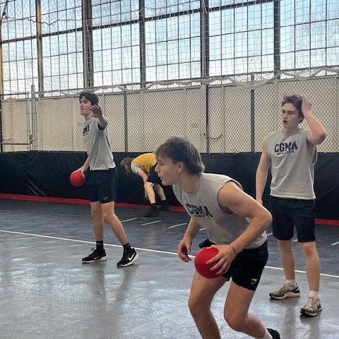 Three boys playing dodgeball in an indoor gym, preparing to throw and dodge a red ball as a girl bends on the floor in the background.