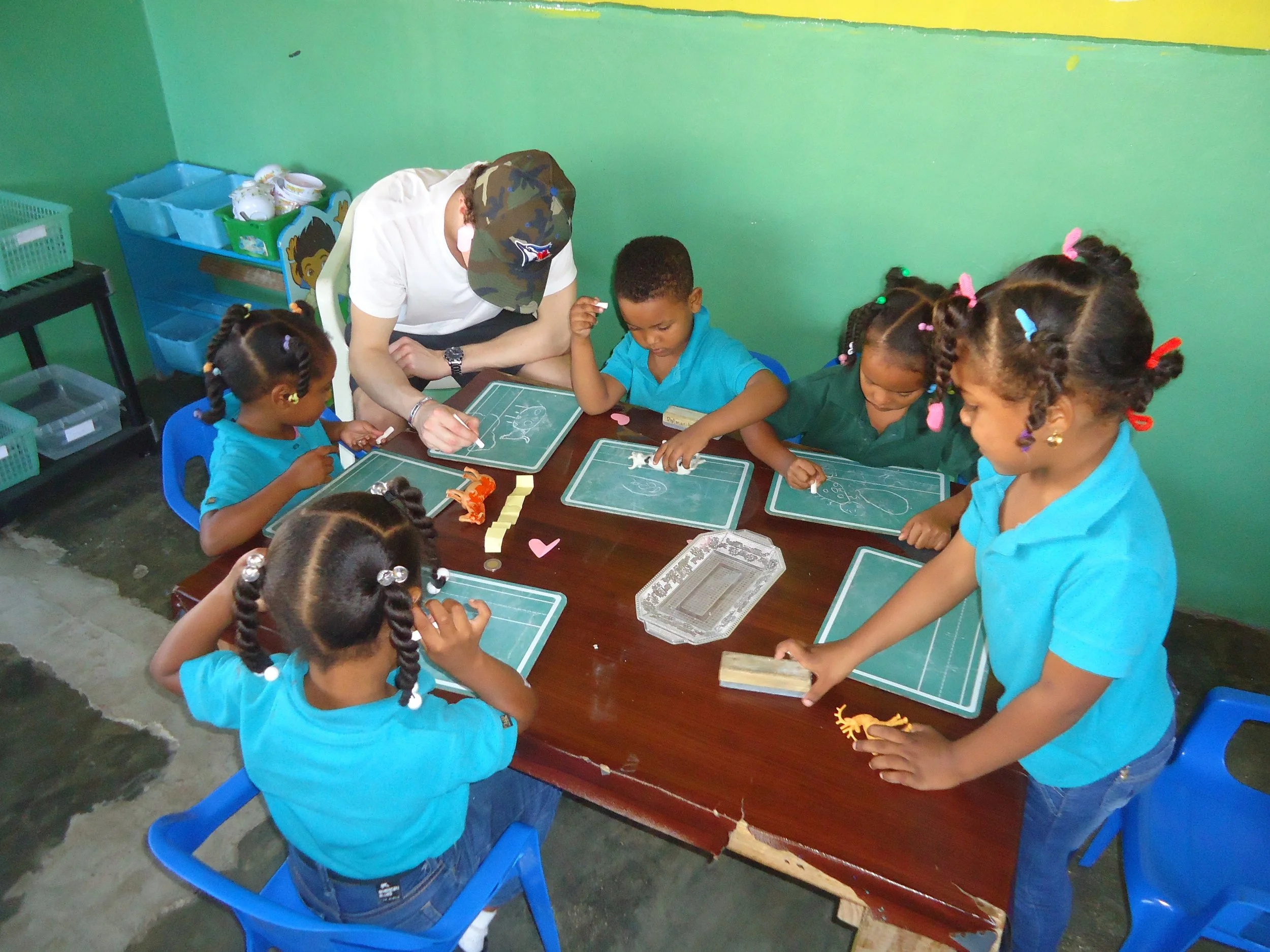 A group of young children with braided hair and colorful hair accessories gathered around a table, working on crafts with a supervising adult in a classroom with green walls.