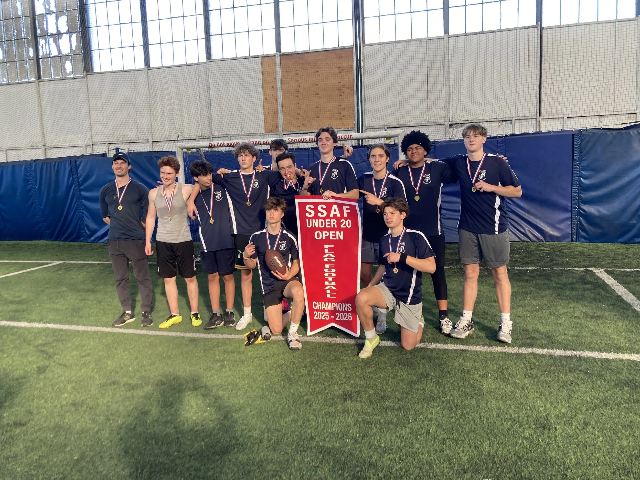A youth football team celebrating their championship victory indoors, wearing medals, holding a trophy, and standing behind a red banner that reads 'SSAF Under 20 Open Champions 2025-2026'.