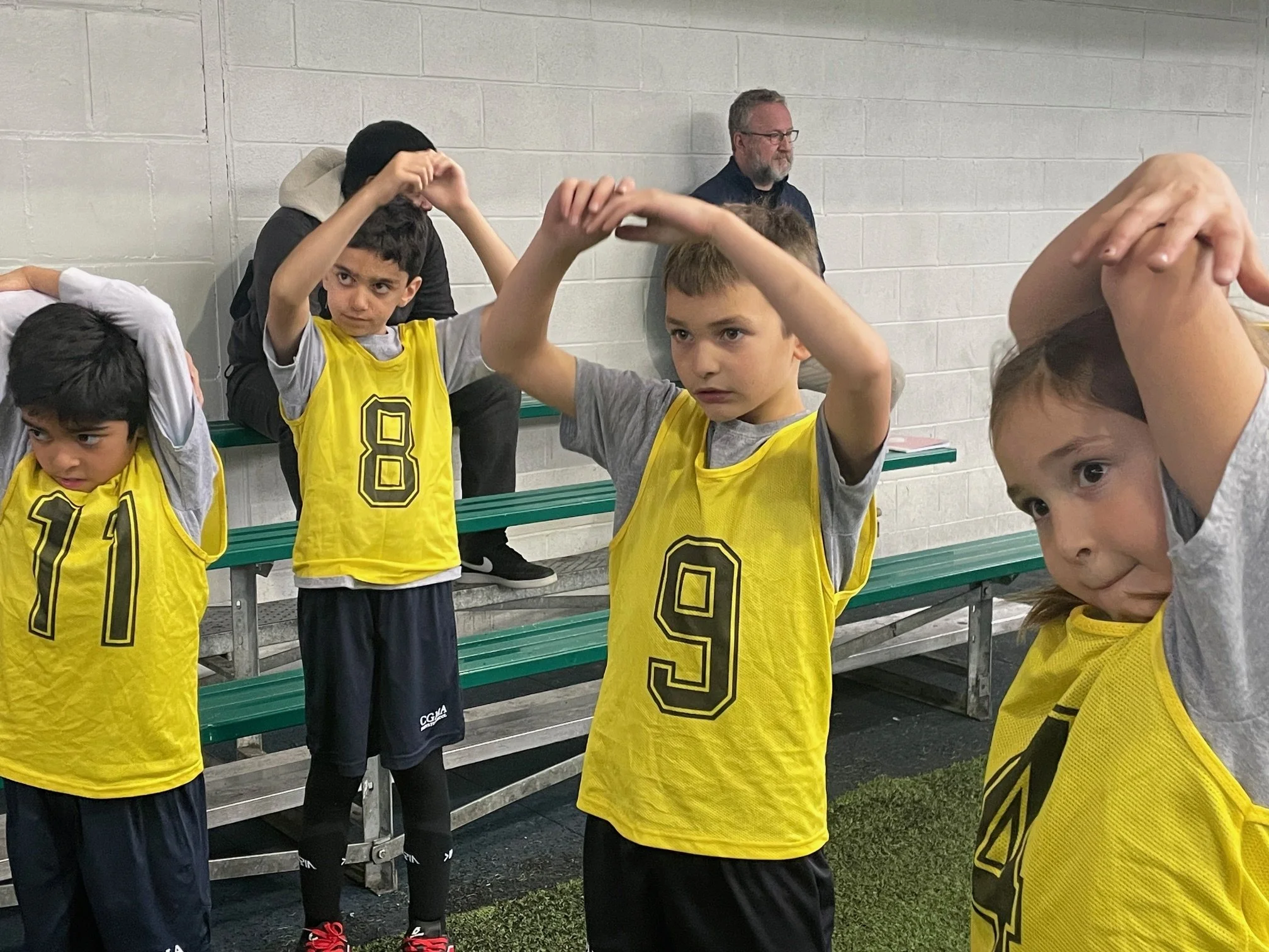 Young boys in yellow sports jerseys stretching while standing against a wall, with an adult man in glasses and a black shirt sitting on a bench in the background.