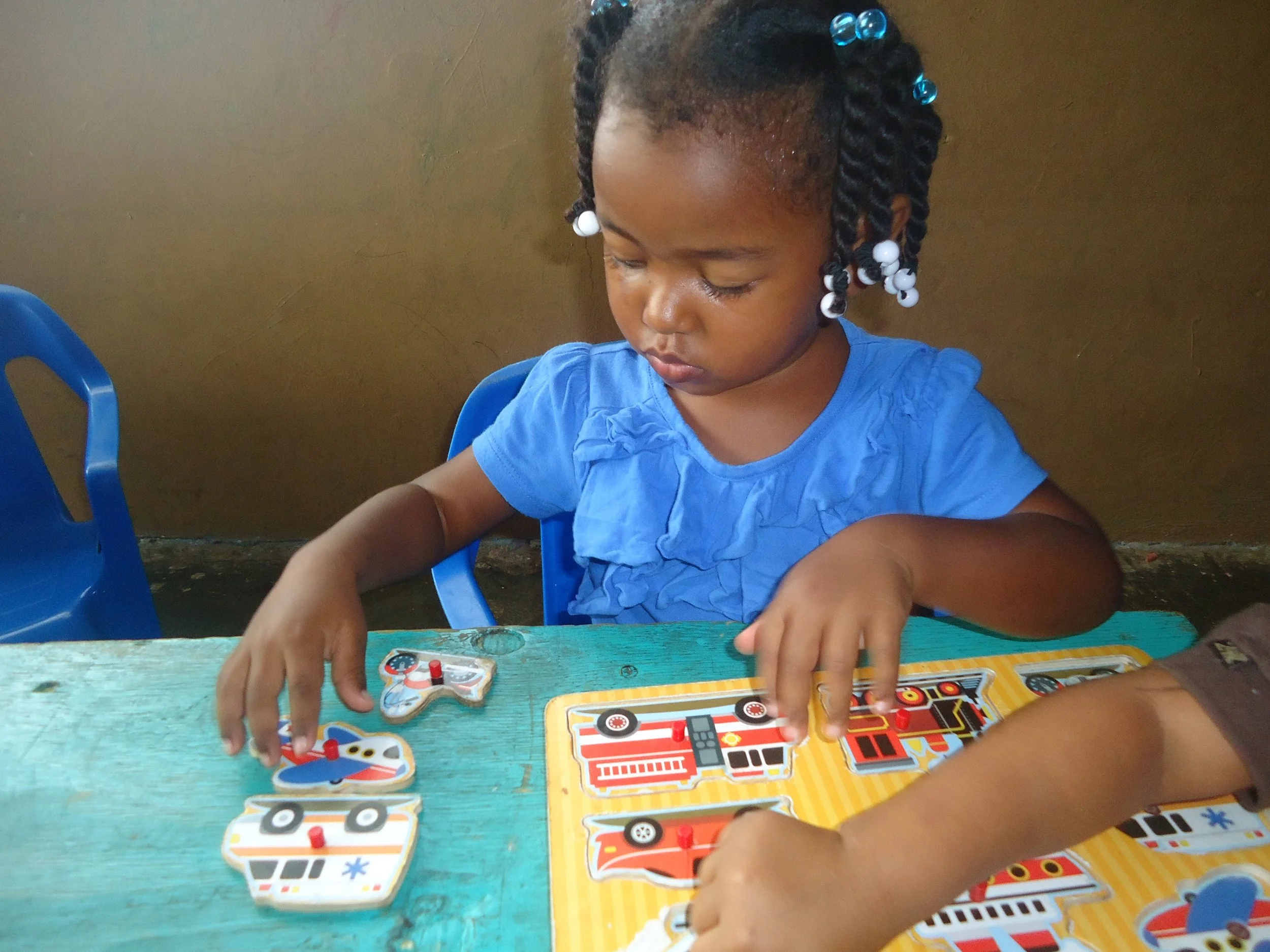 A young girl with braided hair decorated with white and blue beads, focused on playing a board game with colorful illustrations of vehicles and transportation.