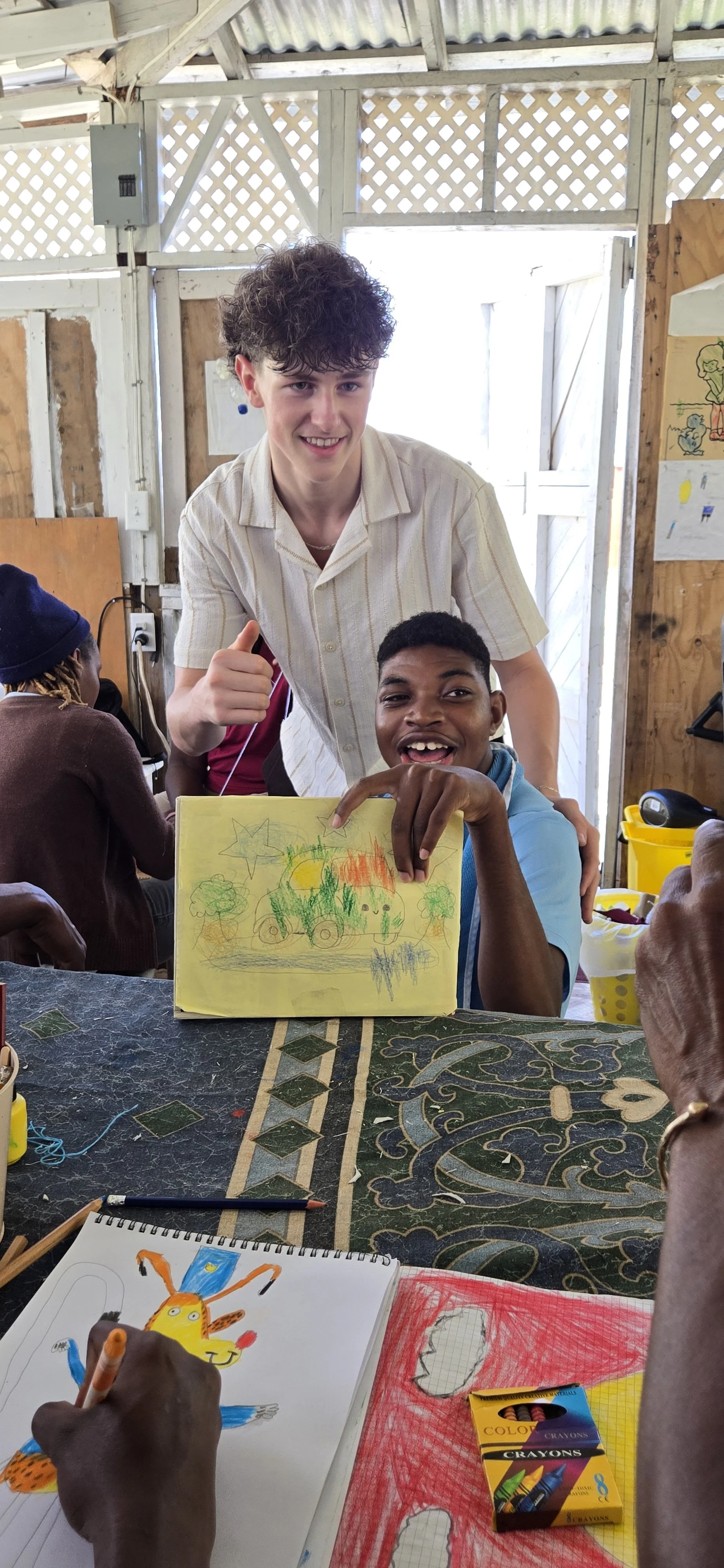 Two boys, one with curly hair and one with short black hair, smile at the camera while sitting at a table with drawings and art supplies. The boy with short hair is holding a drawing of a colorful truck with trees and the sun, and the other boy is wo