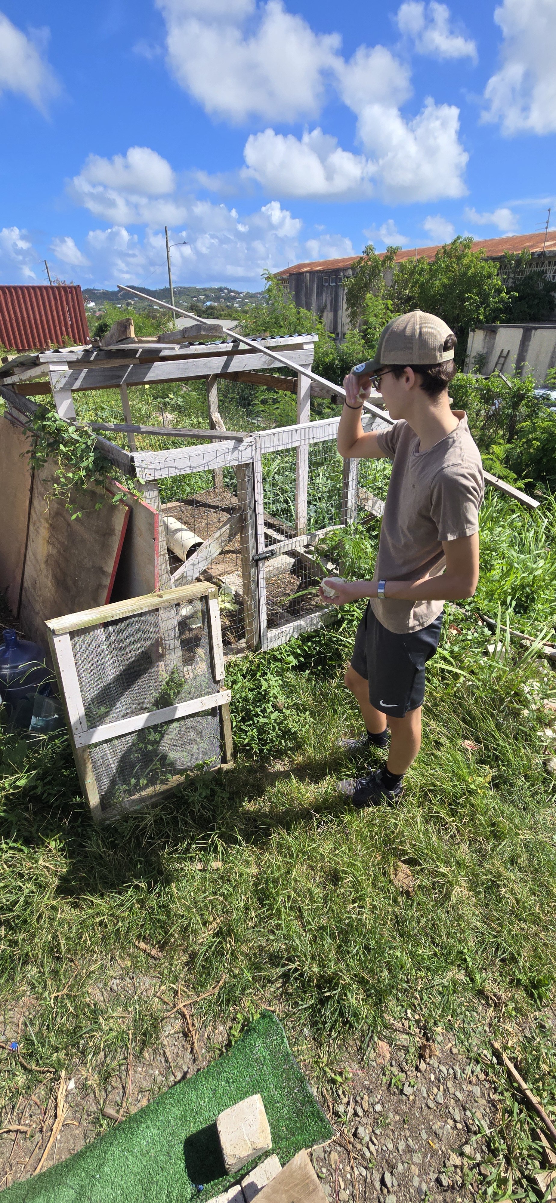 A young man with a tan baseball cap, sunglasses, tan t-shirt, black shorts, and athletic shoes stands outdoors on a grassy area, looking over a small fenced garden with greenery and raised beds, under a partly cloudy blue sky.