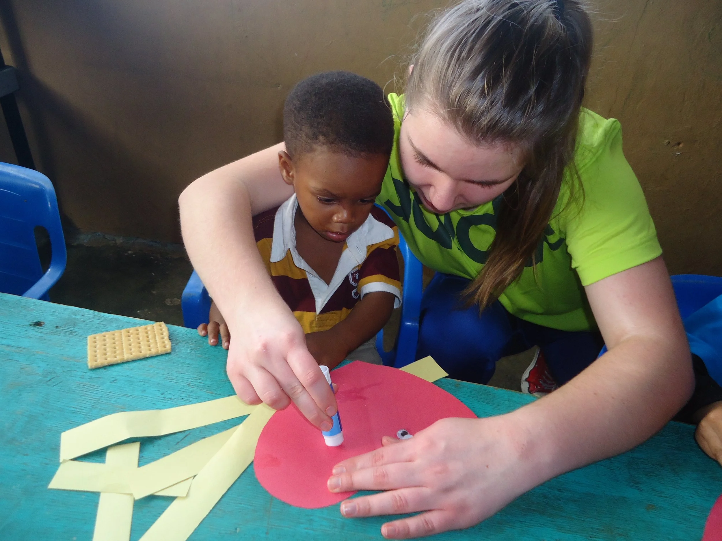 A woman with long brown hair, wearing a bright green shirt, helps a young boy with short black hair, wearing a maroon and yellow striped shirt, with a craft project involving colorful paper and a marker.