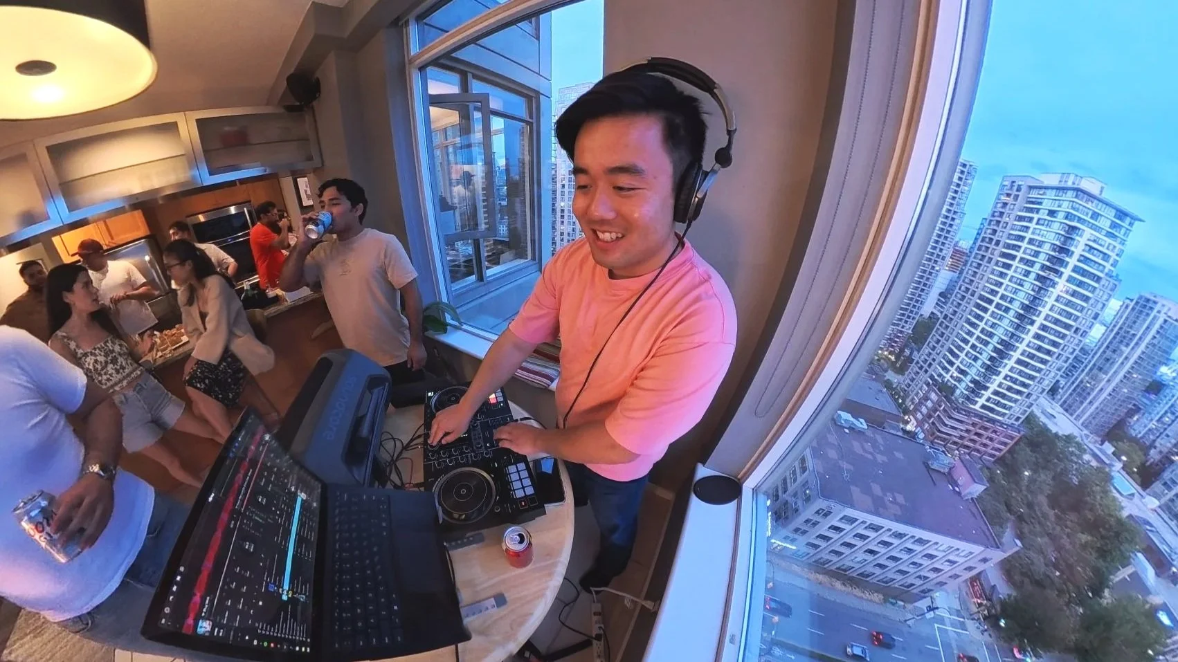 A young man wearing a pink shirt and headphones is DJing at a party inside a high-rise apartment with large windows revealing a cityscape of tall buildings. Several people are socializing in the background.