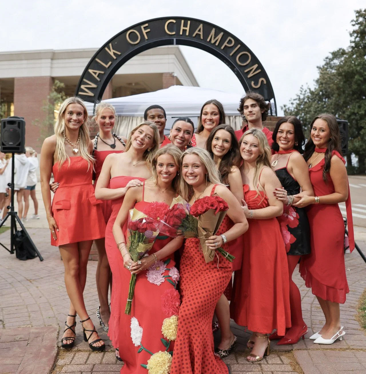Group of people celebrating at a walk of champions event, dressed in red and holding flowers, standing under an arch that says 'Walk of Champions'.
