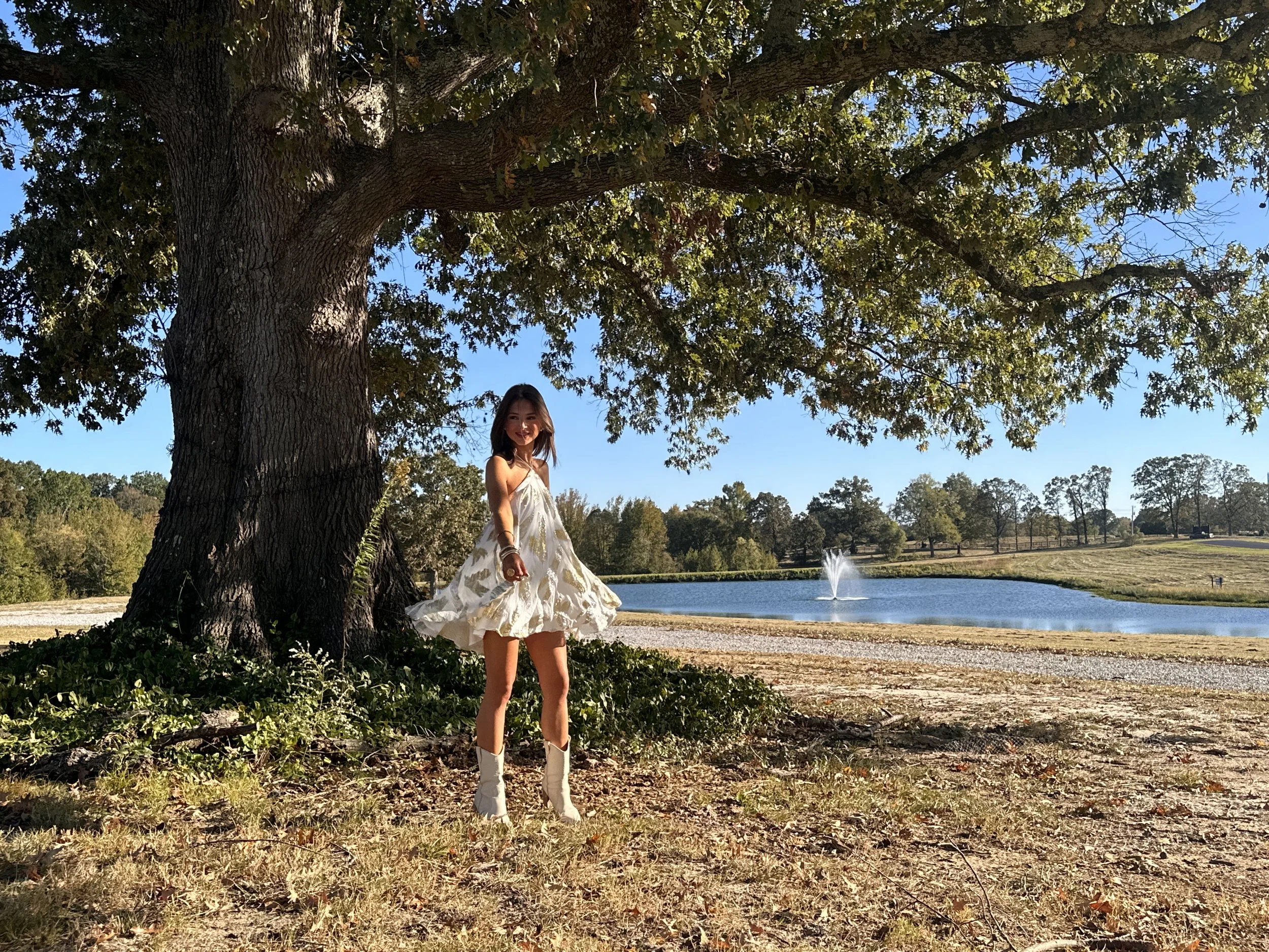 A woman in a white dress and cowboy boots standing outdoors under a large tree near a pond with a fountain, surrounded by a grassy area and more trees in the background.