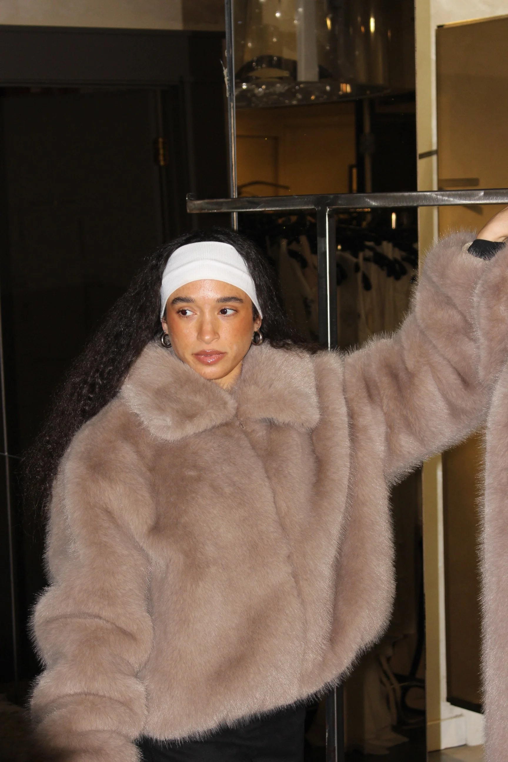 Young woman with long dark hair, wearing a white headband and a beige fur coat, taking a selfie with her arm extended.