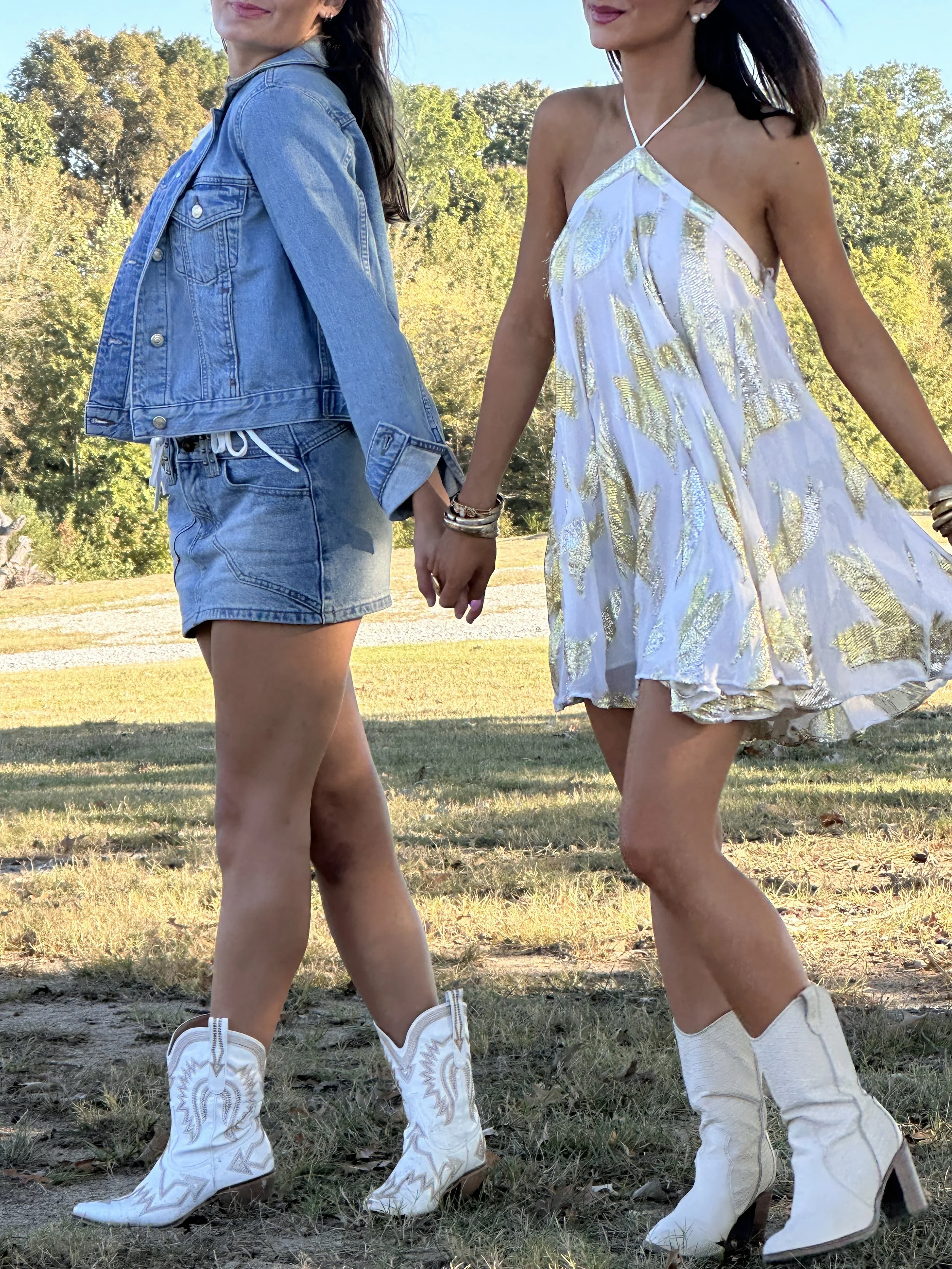Two women holding hands, wearing cowboy boots and Western-style outfits, walking in a park with trees and grass in the background.