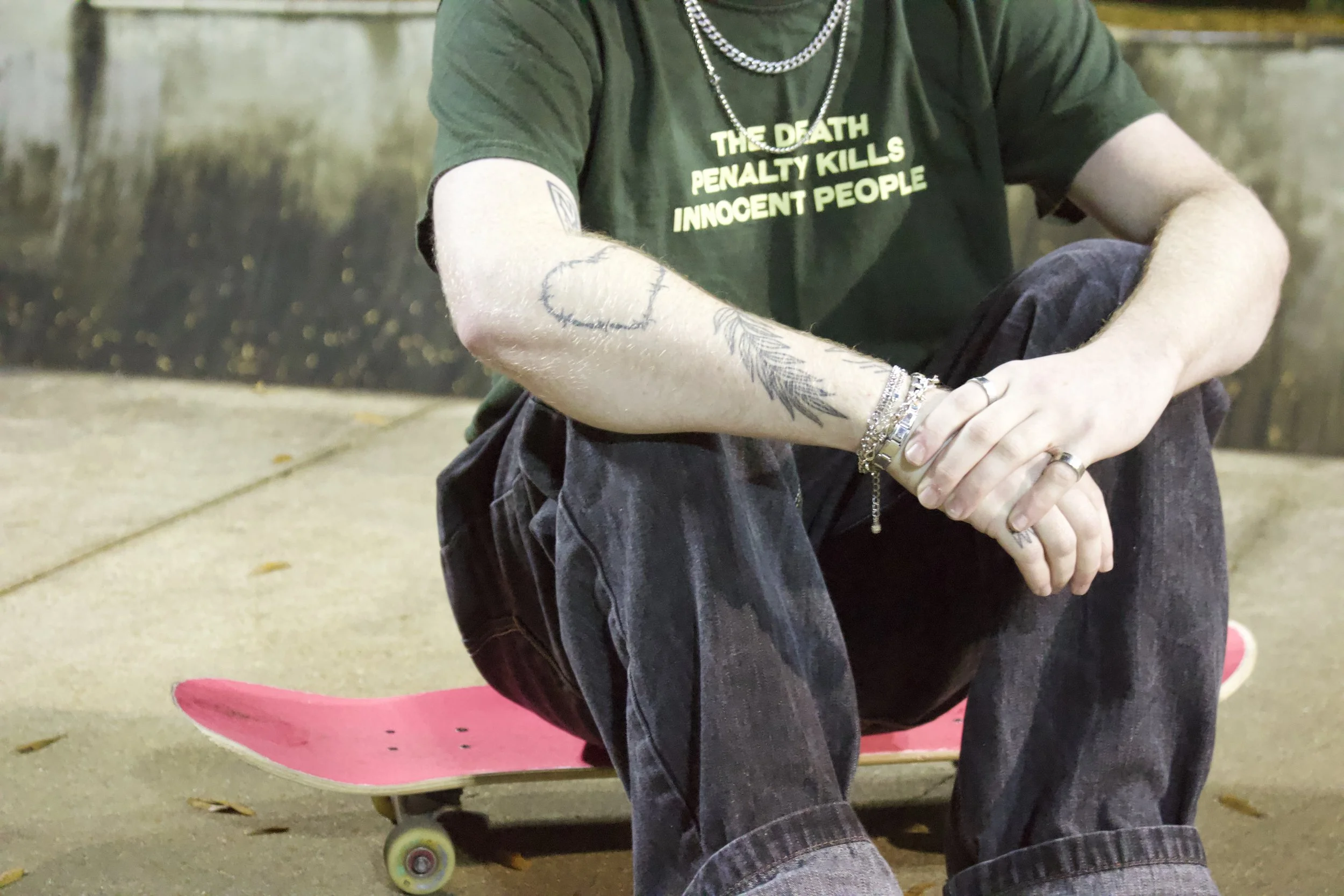 A person sitting with crossed arms on their knees, wearing a black T-shirt with yellow print and silver jewelry, with a skateboard with pink wheels behind them, in an outdoor setting with concrete ground and fallen leaves.