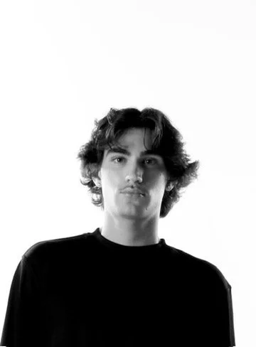 Black and white portrait of a young man with wavy hair wearing a black shirt, against a plain white background.