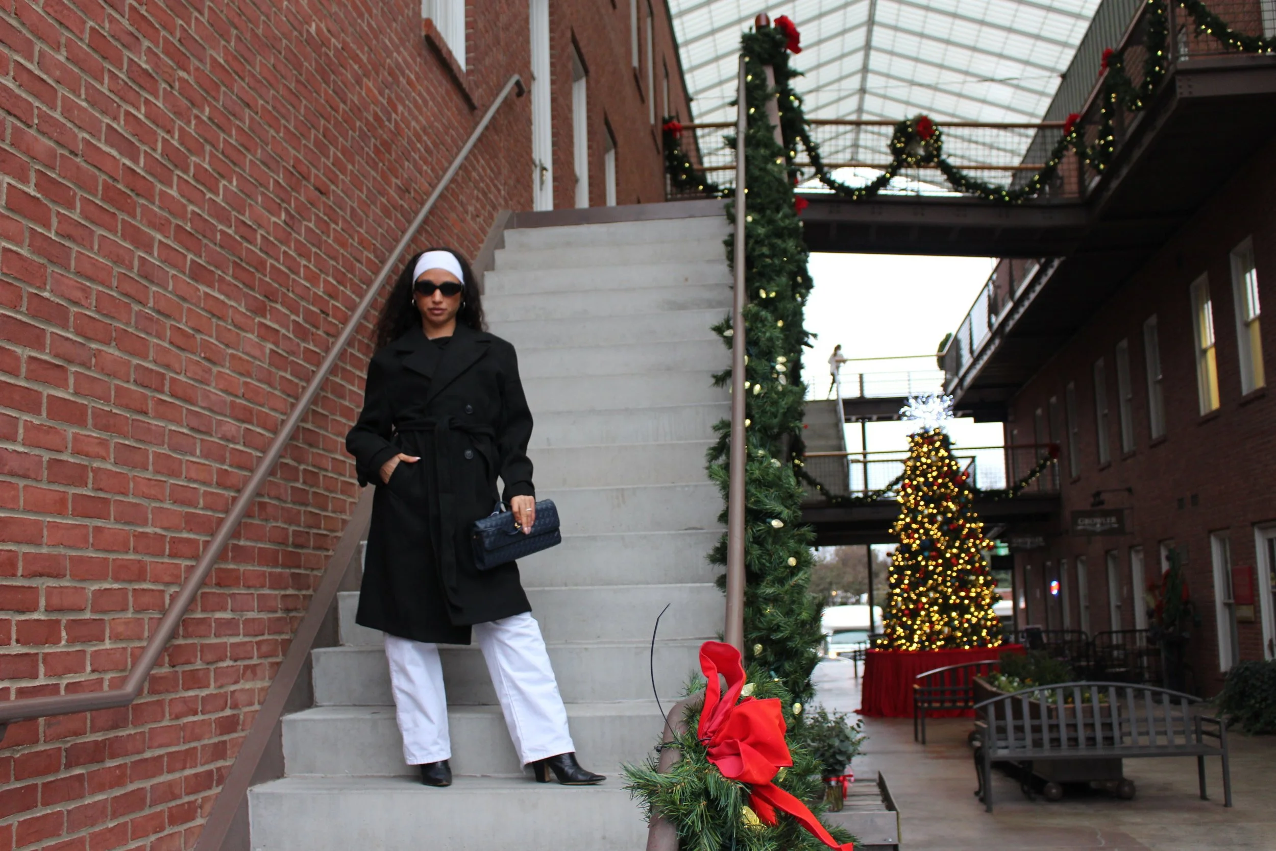A woman dressed in a black coat, white pants, black boots, and sunglasses, standing on a staircase decorated with Christmas garlands and ornaments outside a brick building with Christmas trees in the background.