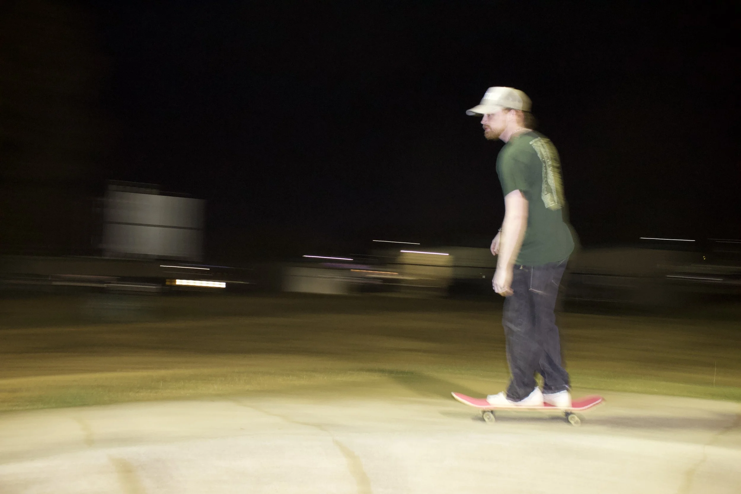 A person skateboarding at night, wearing a beige cap, green t-shirt, dark jeans, and white shoes, on a pink skateboard, with a dark background and blurred lights.