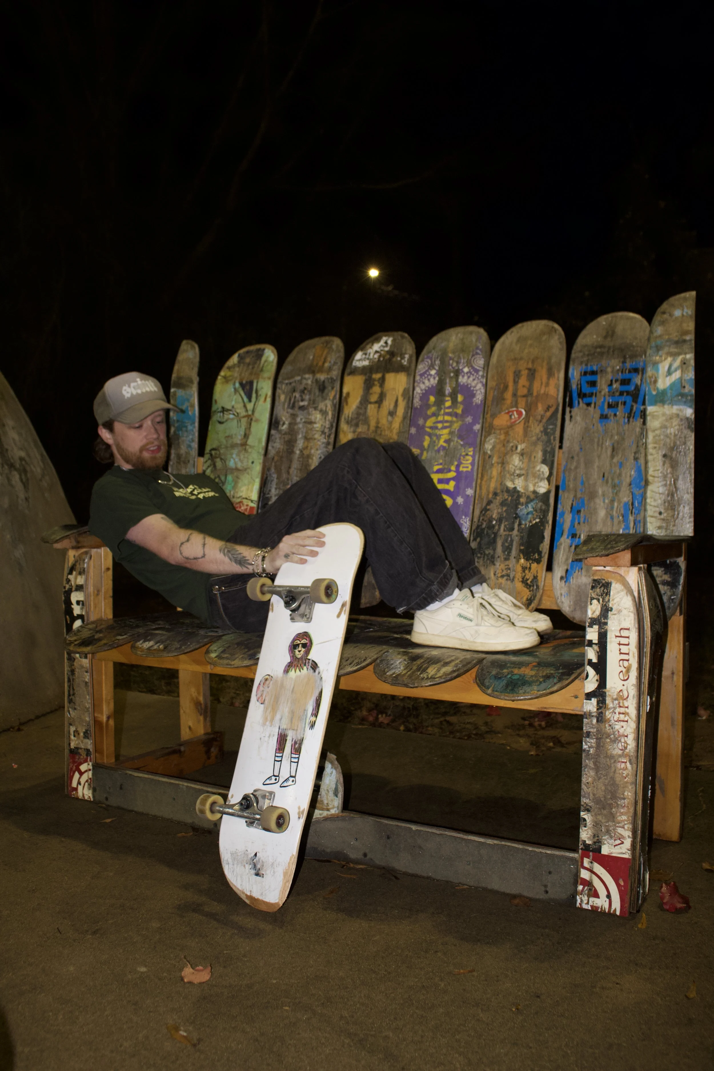 A man relaxing on a skateboard bench at night, with skateboards with graffiti and artwork behind him and a moon in the night sky.