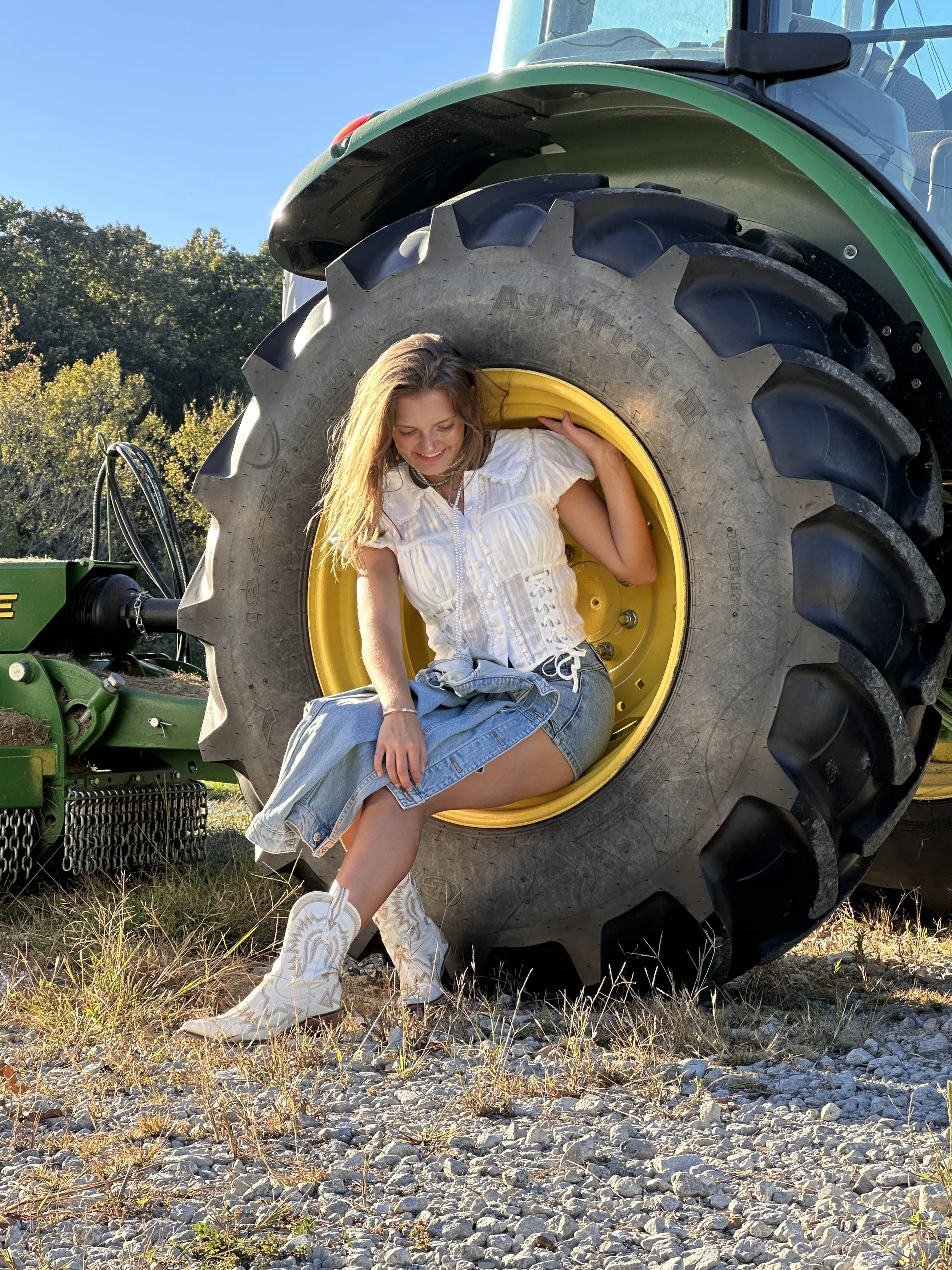 A young woman sitting inside the large tire of a green tractor outdoors on a sunny day, smiling and looking down, with trees and a clear blue sky in the background.