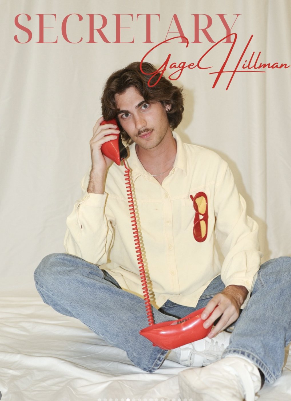 Book cover titled 'Secretary' by Gage Hillman, featuring a young man with brown hair and a mustache, sitting cross-legged on a white surface, holding a red telephone receiver to his ear, with red sunglasses clipped to his shirt pocket, against a plai
