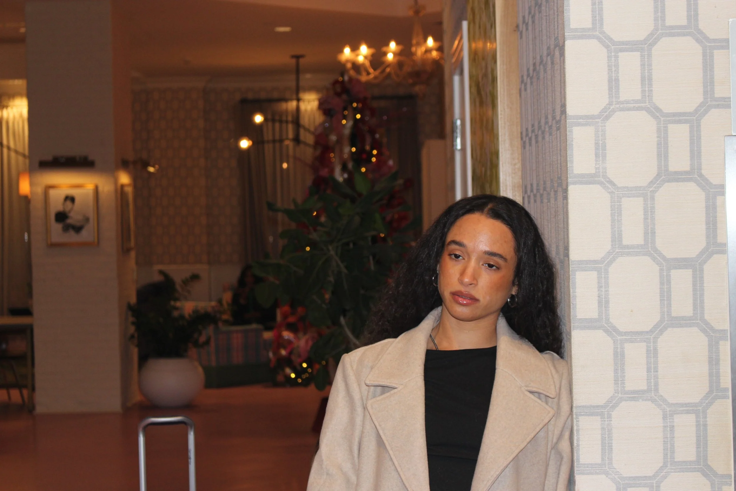 A woman with curly dark hair wearing a beige coat and black top, standing indoors near a patterned wall with a Christmas tree in the background decorated with lights and ornaments.