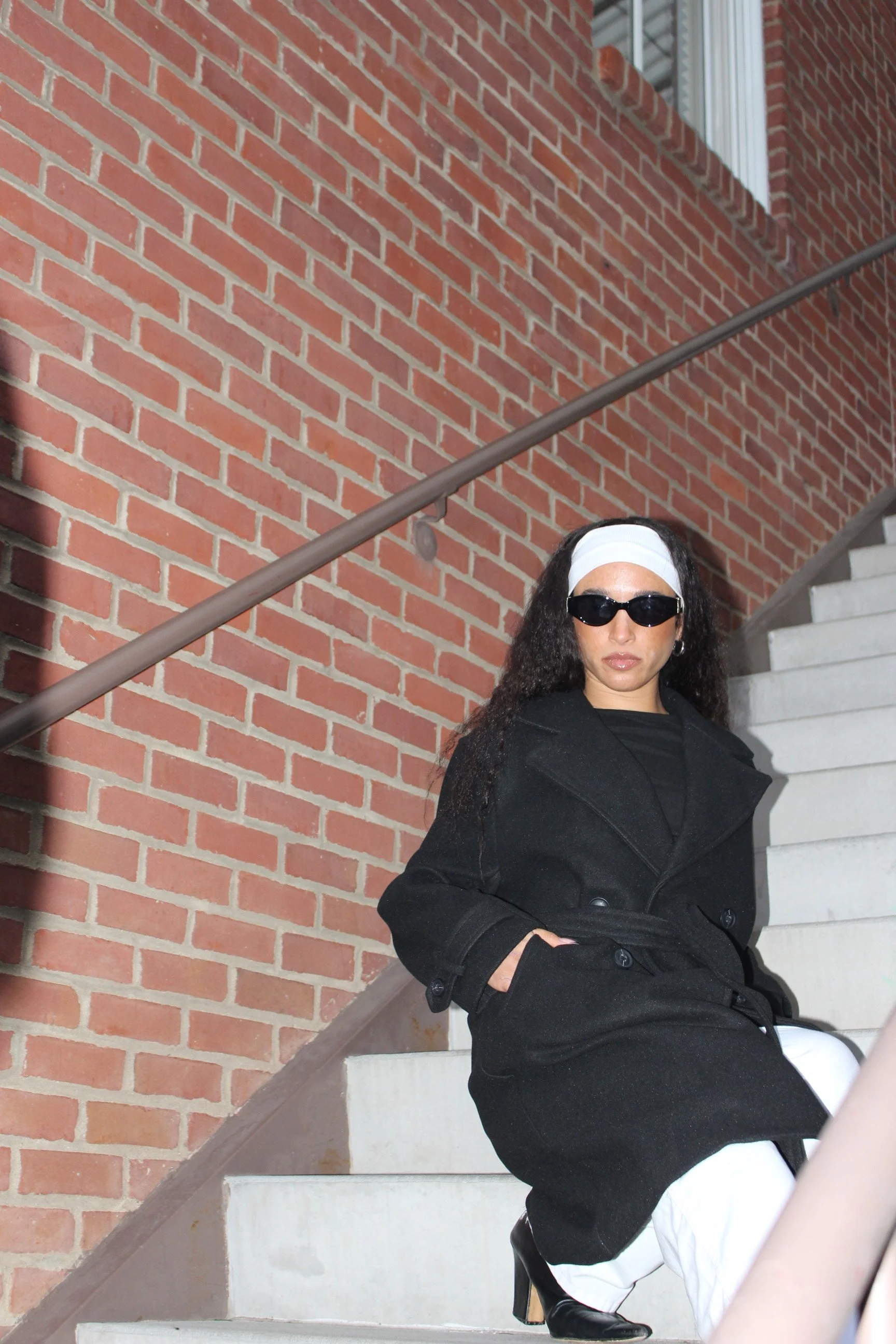 A woman sitting on outdoor concrete stairs, wearing a black coat, white pants, black heels, sunglasses, and a white headband, with a brick wall and railing in the background.