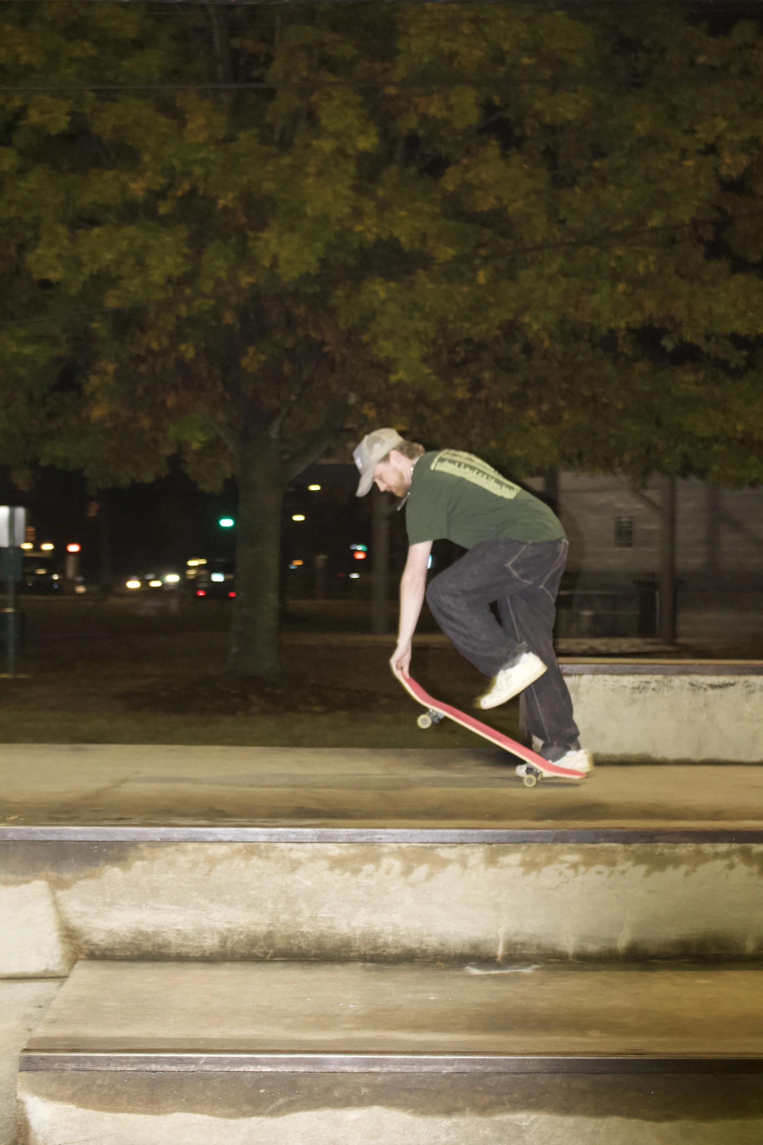 A person skating on a curb at night, wearing a green shirt and gray pants, with trees and streetlights in the background.