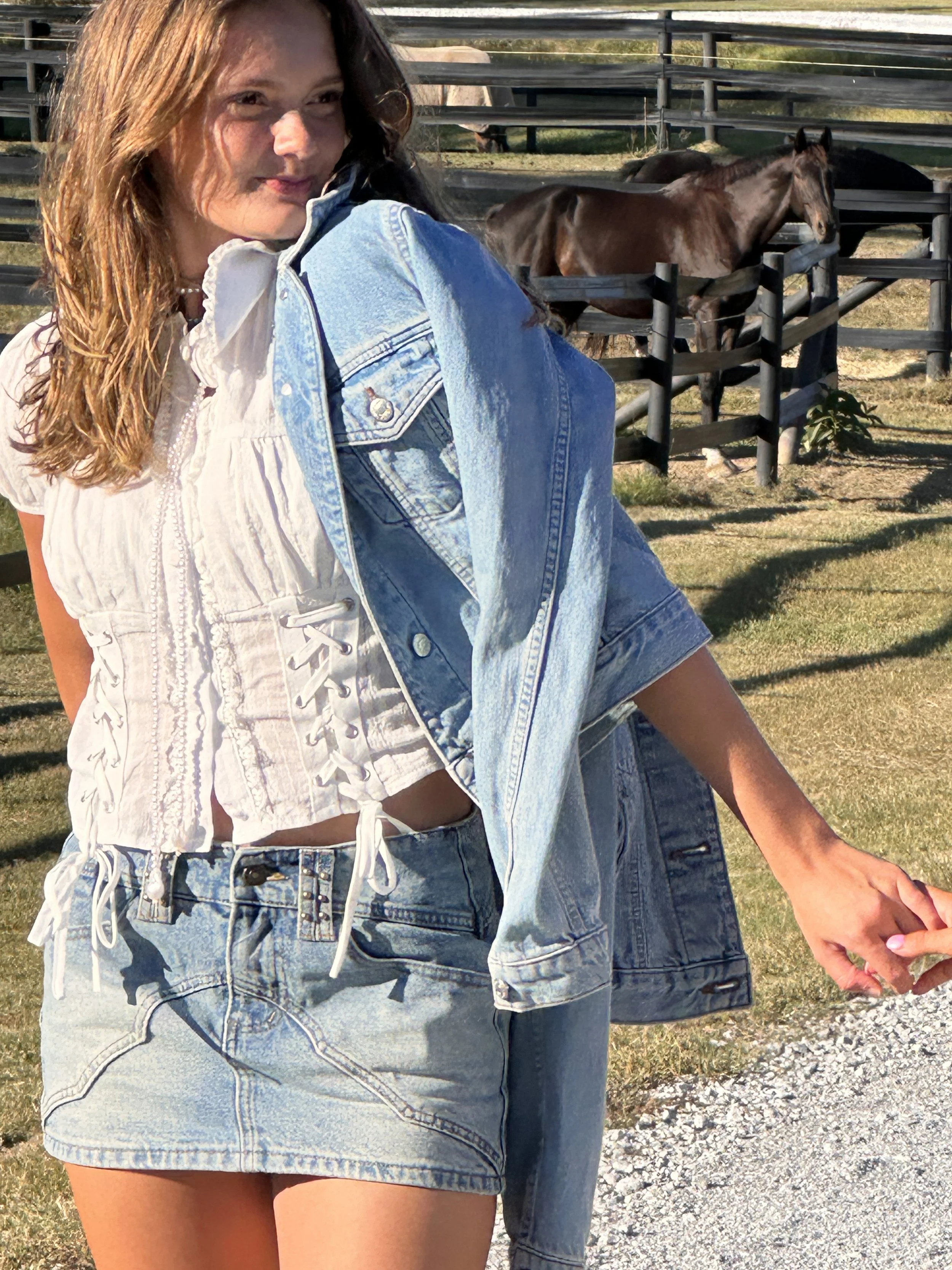 A woman with red hair wearing a denim jacket and shorts, standing outdoors near a fenced area with horses.