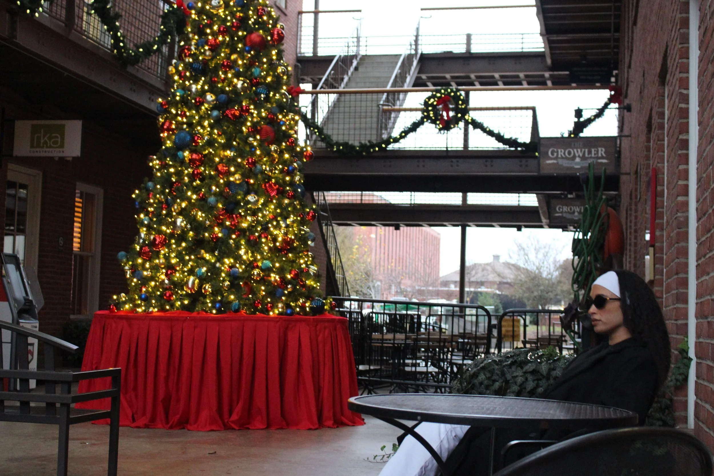 A decorated Christmas tree with ornaments and lights, placed on a red-covered table in an outdoor area with brick buildings, a fire escape, and holiday decorations in the background, during daytime.