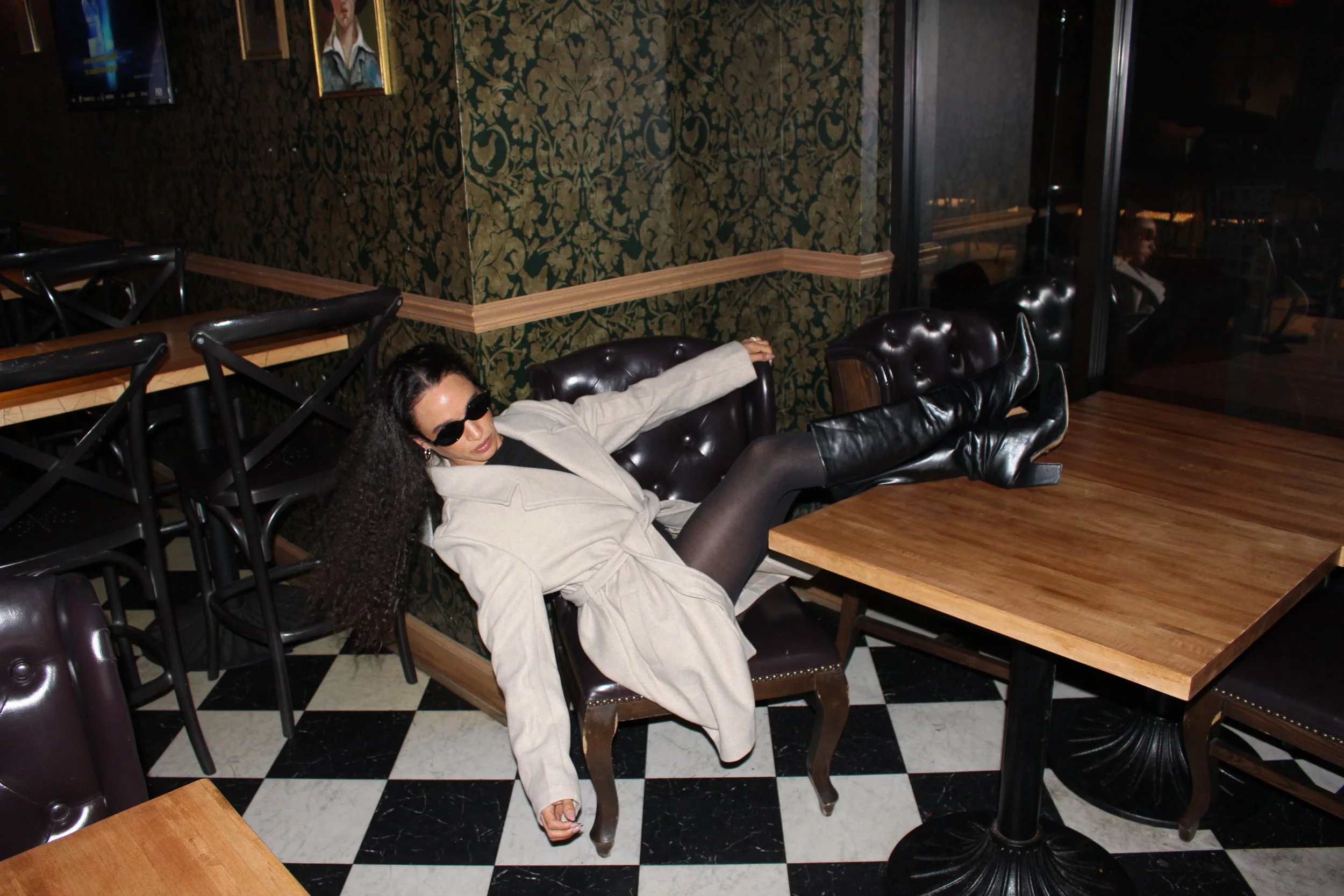 Woman with long curly hair wearing sunglasses, a light-colored coat, and black leather boots relaxing on a leather armchair in a dimly lit restaurant with patterned wallpaper and checkered floor.