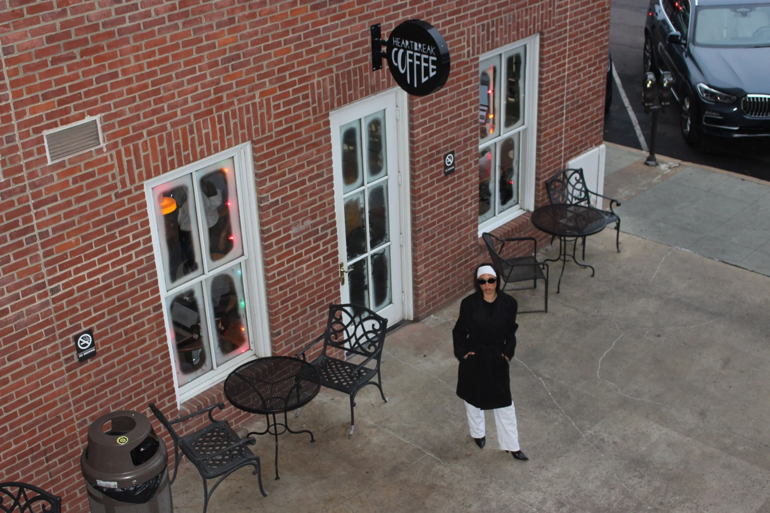 A woman in black jacket and white pants standing outside a brick coffee shop called Heartbreak Coffee, with tables and chairs around, parking lot with a black car, and a woman with sunglasses and white head covering in the foreground.
