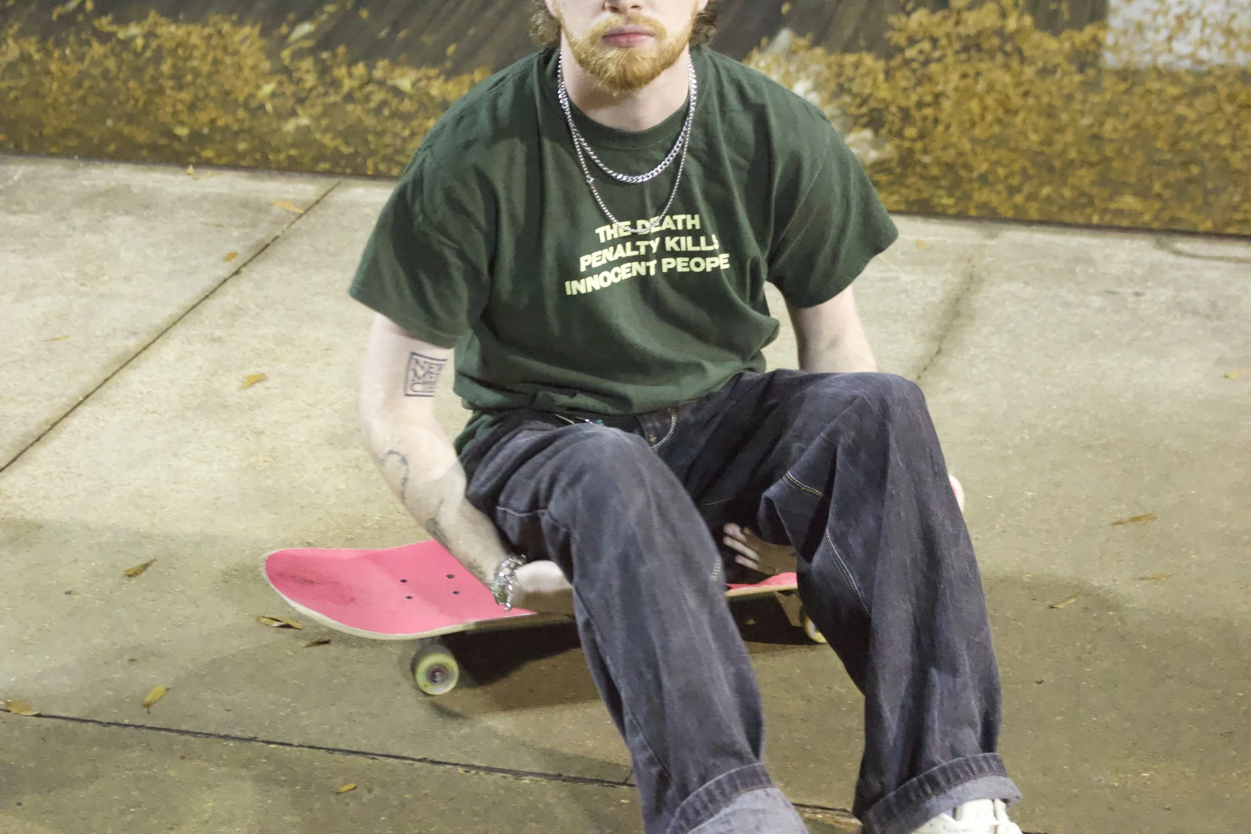 A person with a beard and tattoos wearing a black t-shirt with green text, dark pants, and white shoes sitting on a skateboard at night on a concrete sidewalk with fallen leaves and a mural in the background.